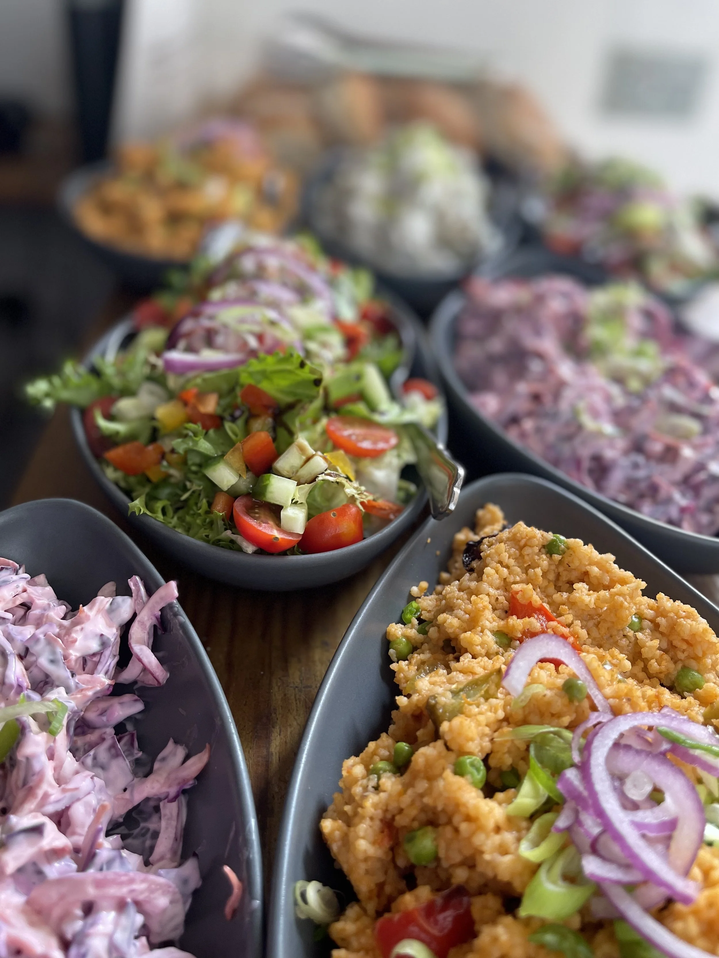 Assorted bowls of colorful salads and side dishes on a wooden table, featuring vegetables, pasta, and creamy dressings.