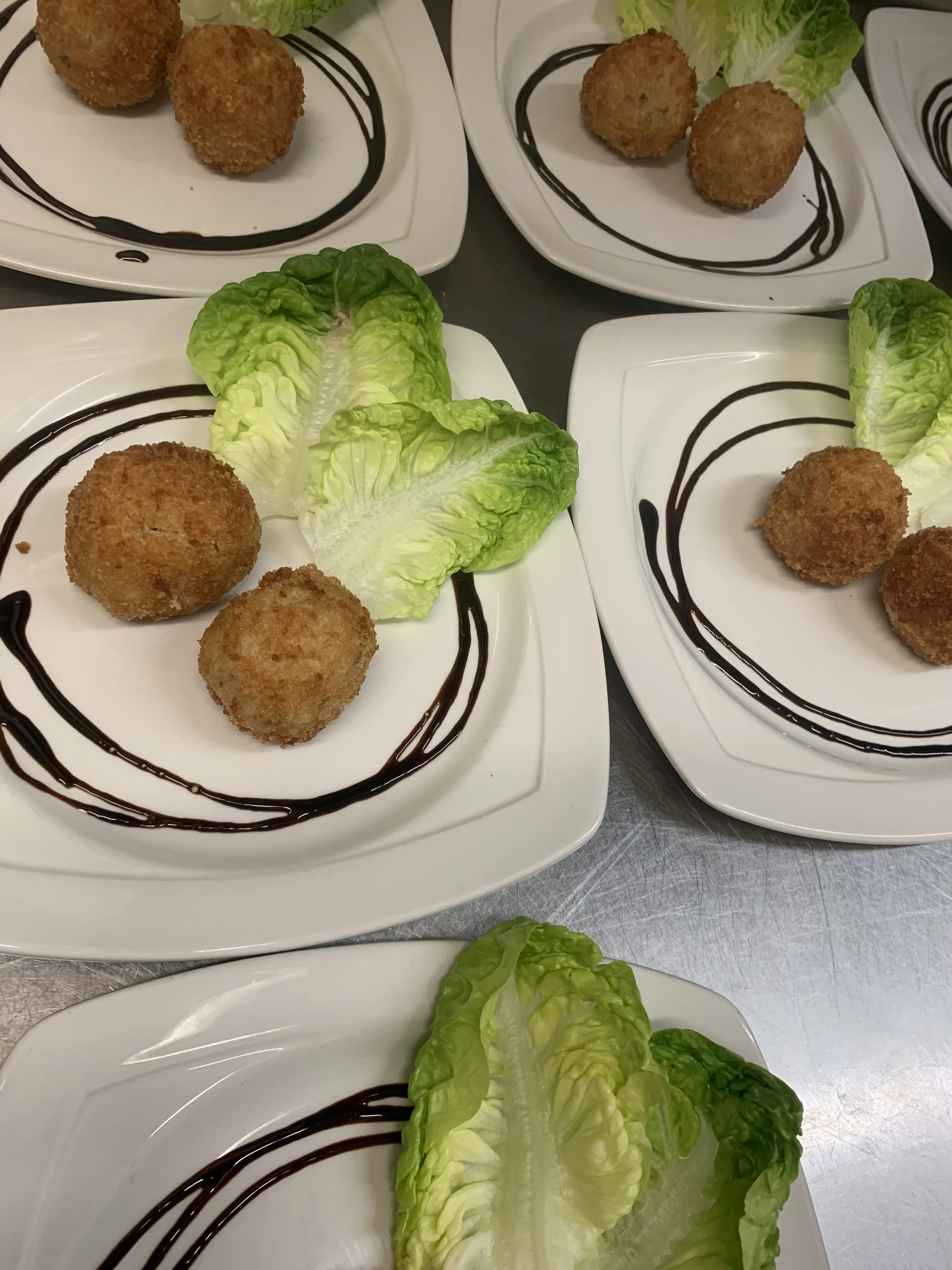 Four white plates with three breaded and fried spherical appetizers, garnished with drizzle of chocolate sauce, and a romaine lettuce leaf on each plate.