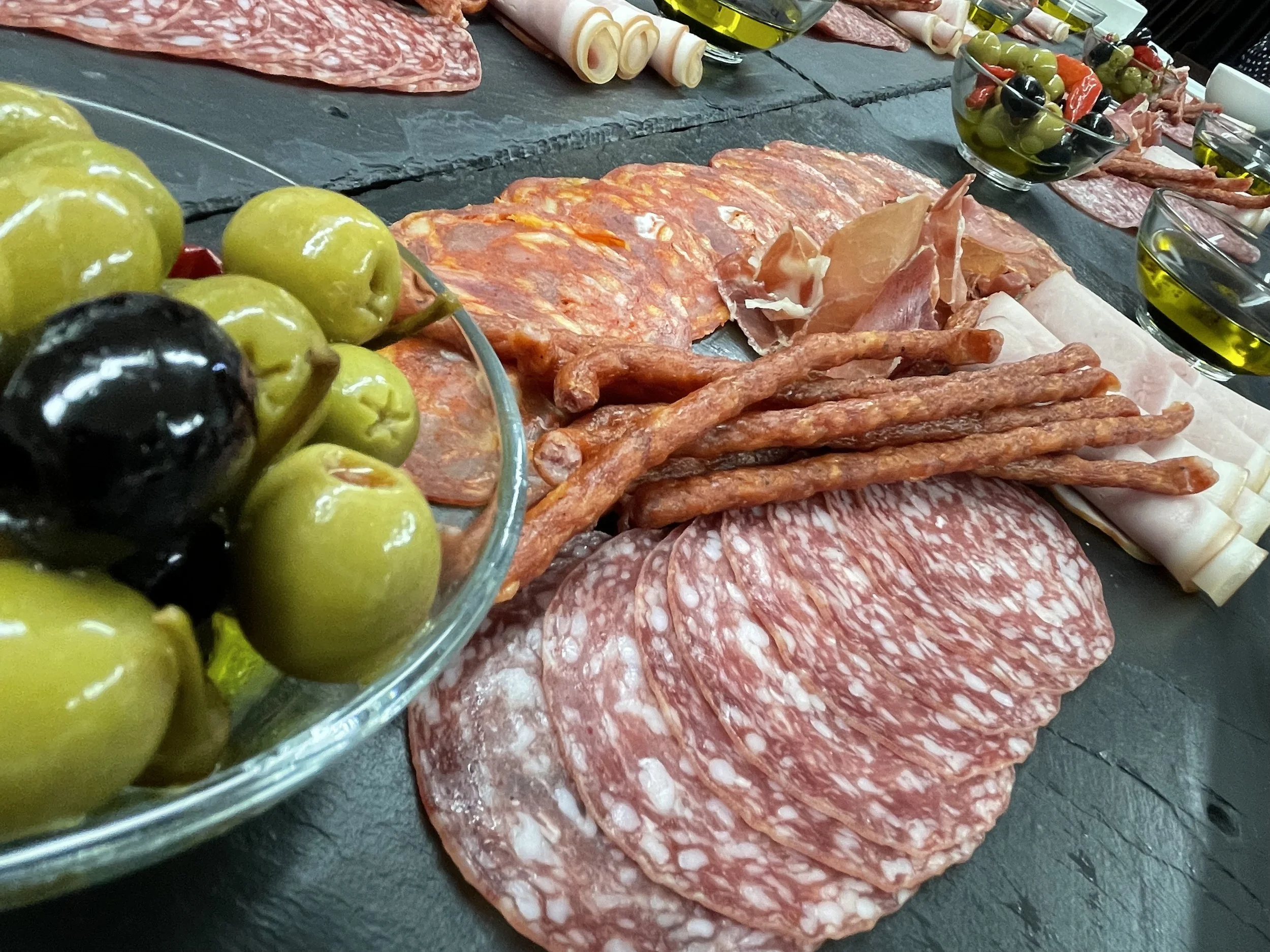 Assorted cured meats, green and black olives, and bowls of olive oil on a dark slate serving platter.