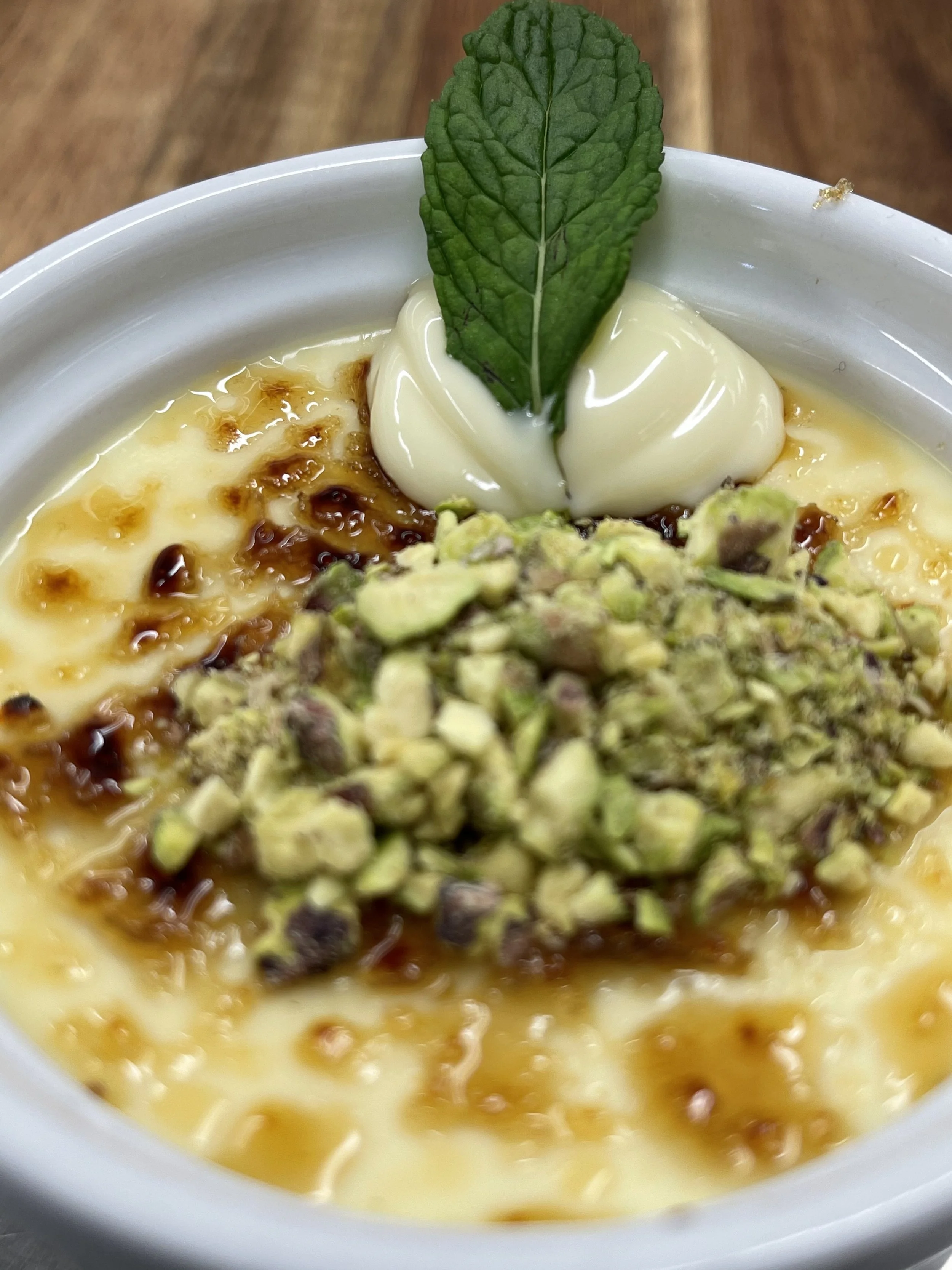 Close-up of a dessert with a topping of chopped pistachios, a dollop of cream with a mint leaf, and caramelized sugar.