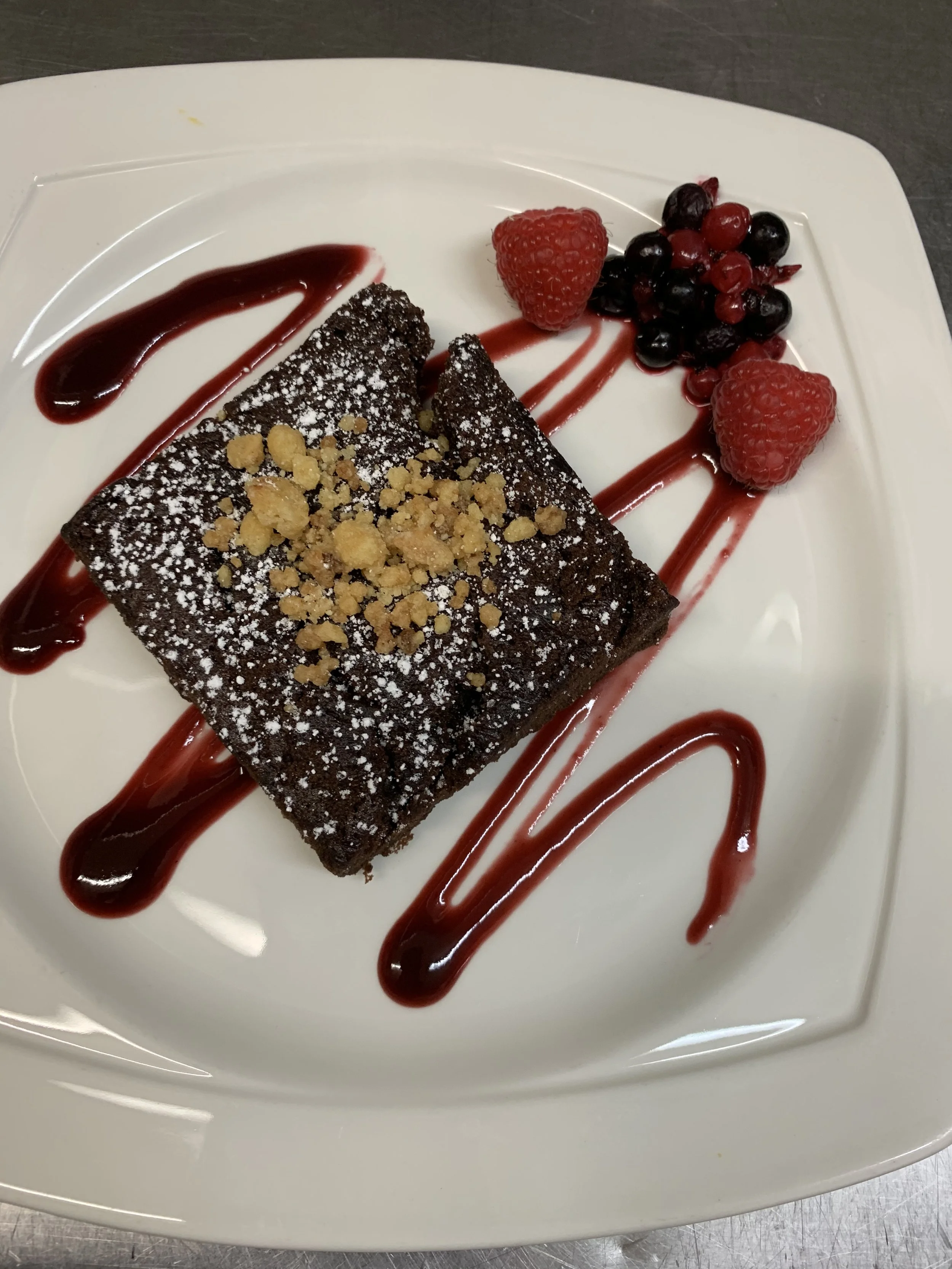 Chocolate brownie dessert garnished with crushed nuts and powdered sugar, served with raspberry and mixed berries, drizzled with berry sauce on a white plate.