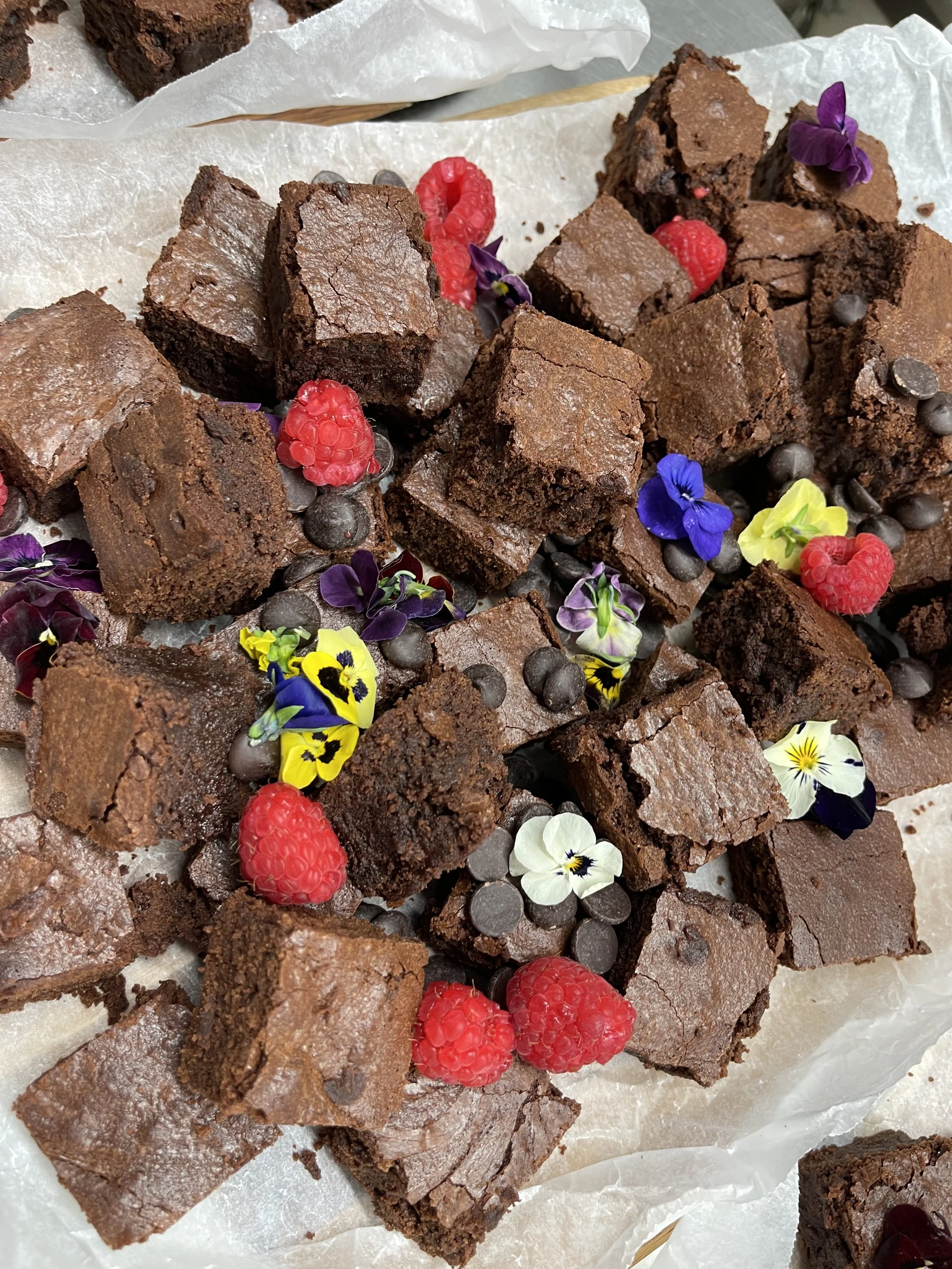 Close-up of a assortment of brownie squares topped with fresh raspberries, edible flowers, and chocolate chips on parchment paper.