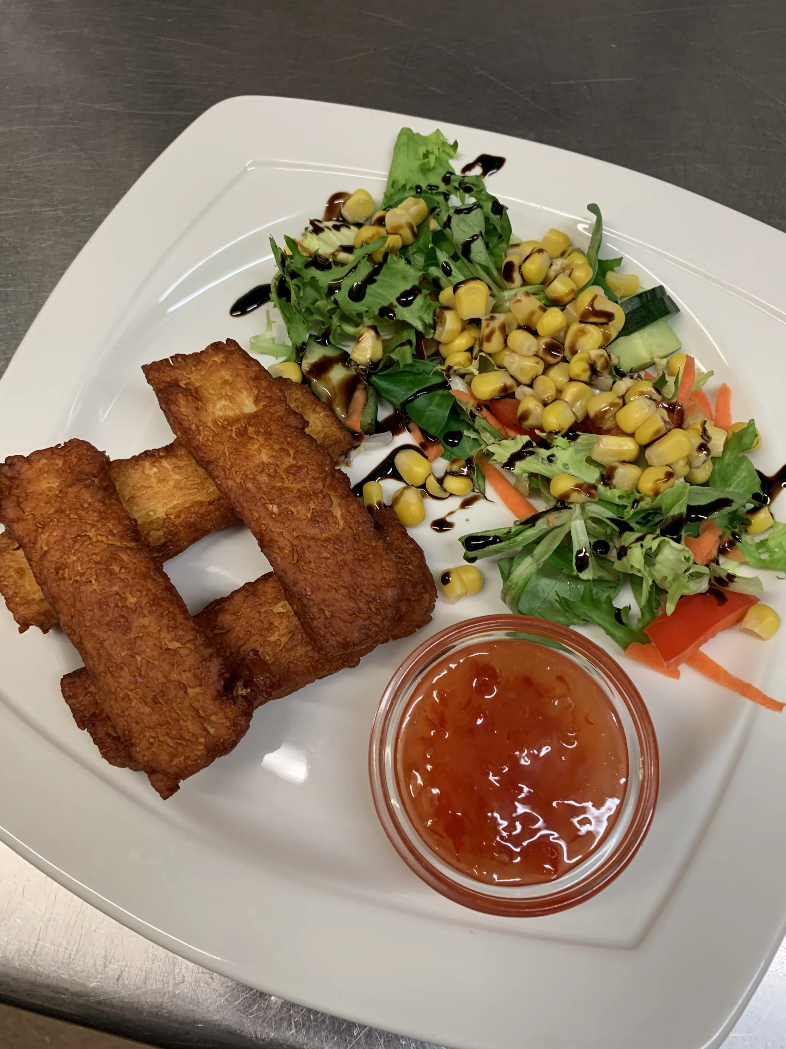Fried fish sticks with a side salad of mixed greens, corn, cucumber, and shredded carrots, served with a small bowl of orange sauce or salsa.