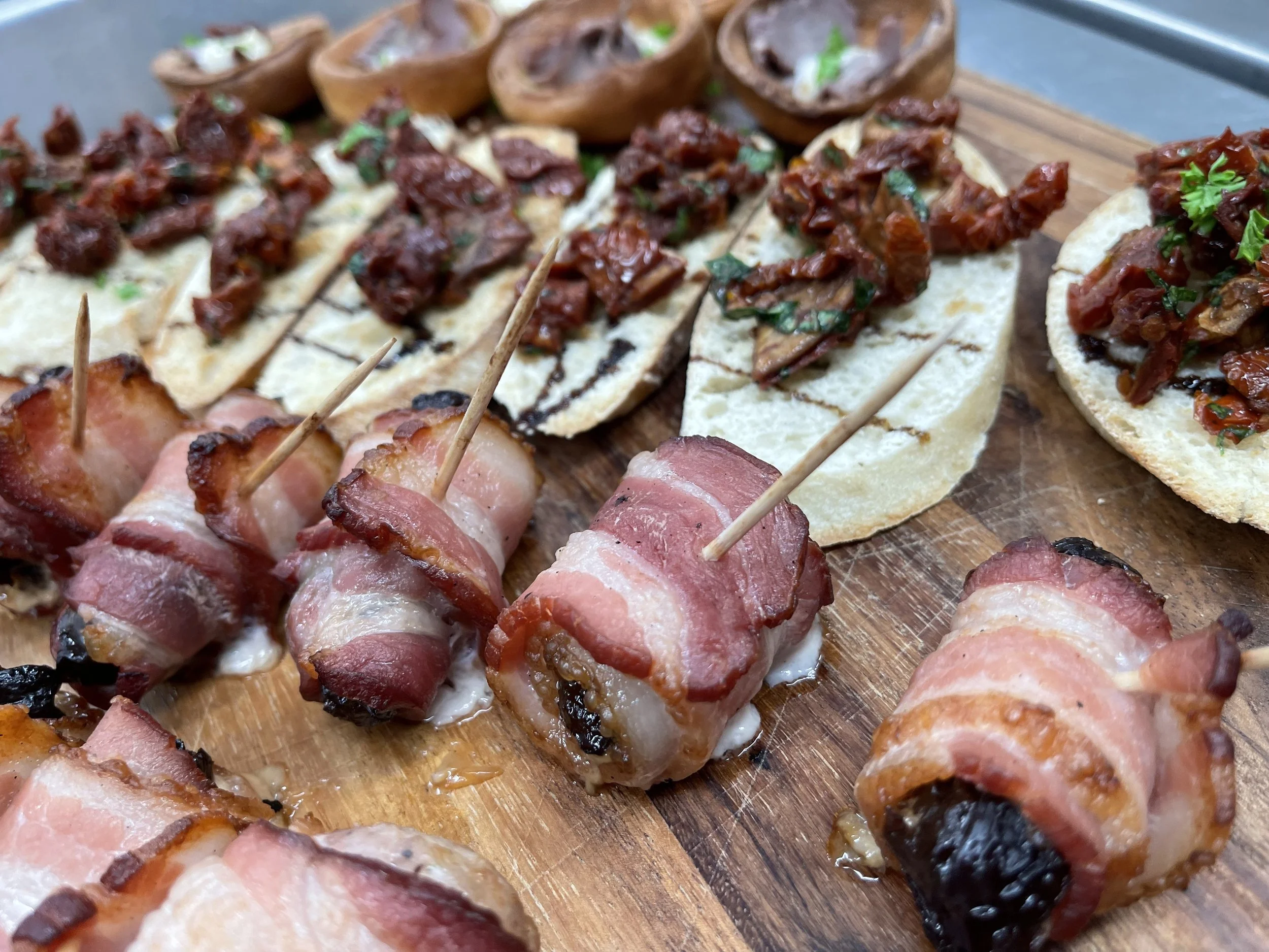 Assorted appetizers on a wooden serving board, including bacon-wrapped items and mini toasts topped with sun-dried tomatoes and herbs.
