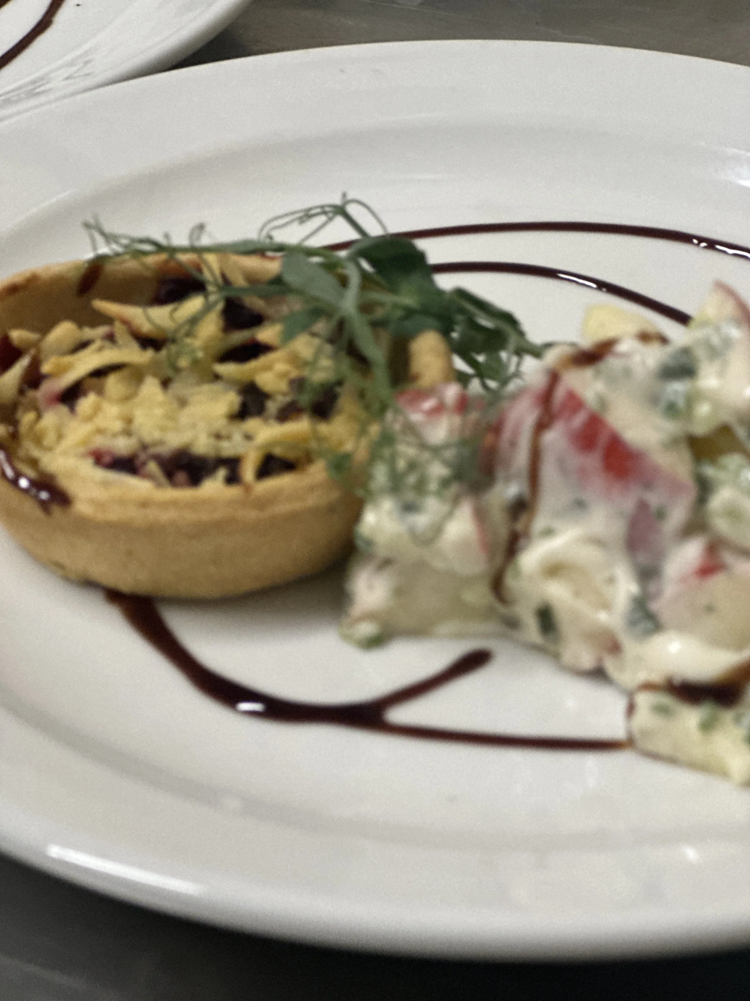 A dessert with a halved tart filled with cream, topped with shredded chocolate and garnished with a sprig of herbs, served with a side of fruit salad on a white plate decorated with chocolate syrup.