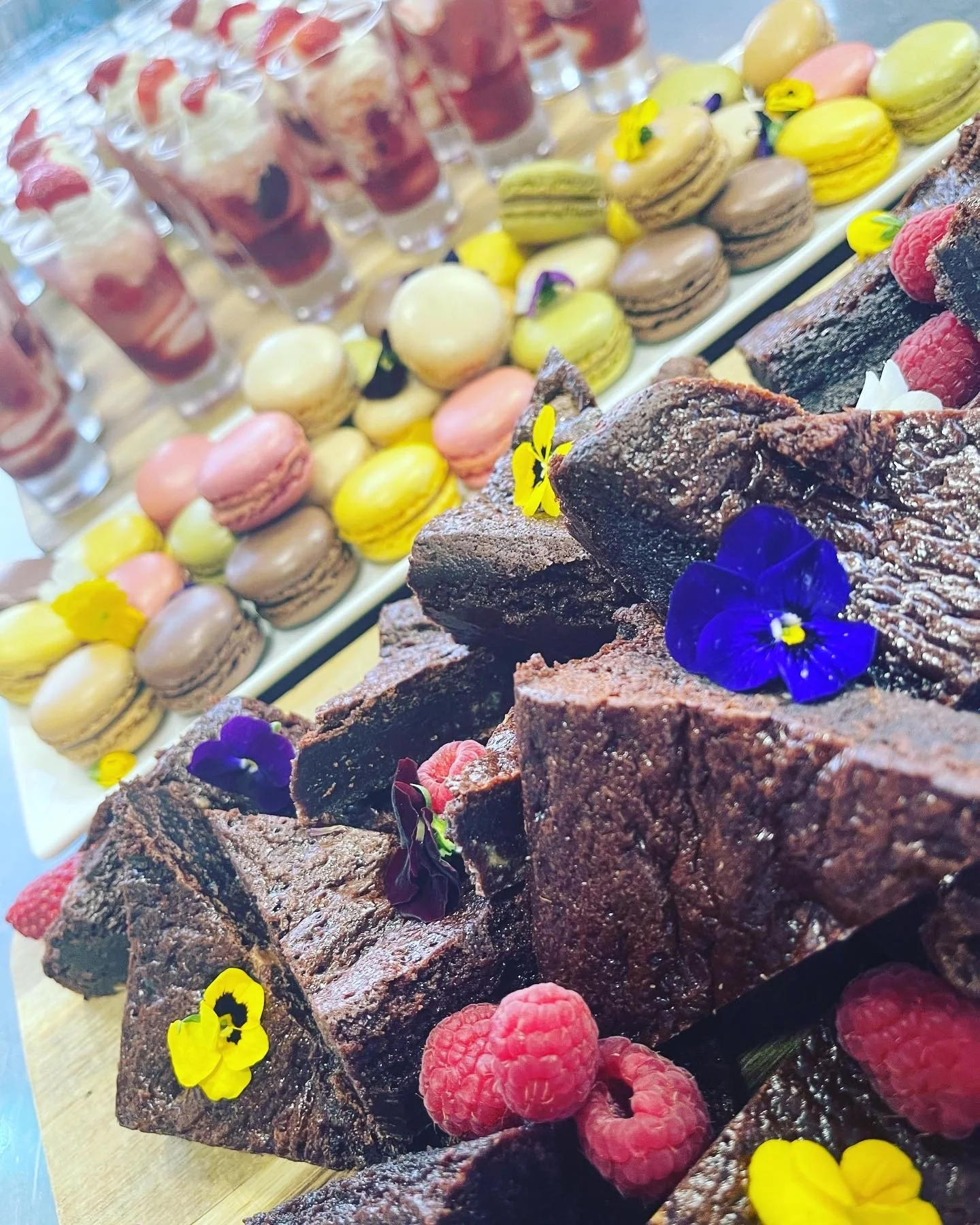 Assorted desserts including brownies with edible flowers and raspberries, and macarons in pastel colors.
