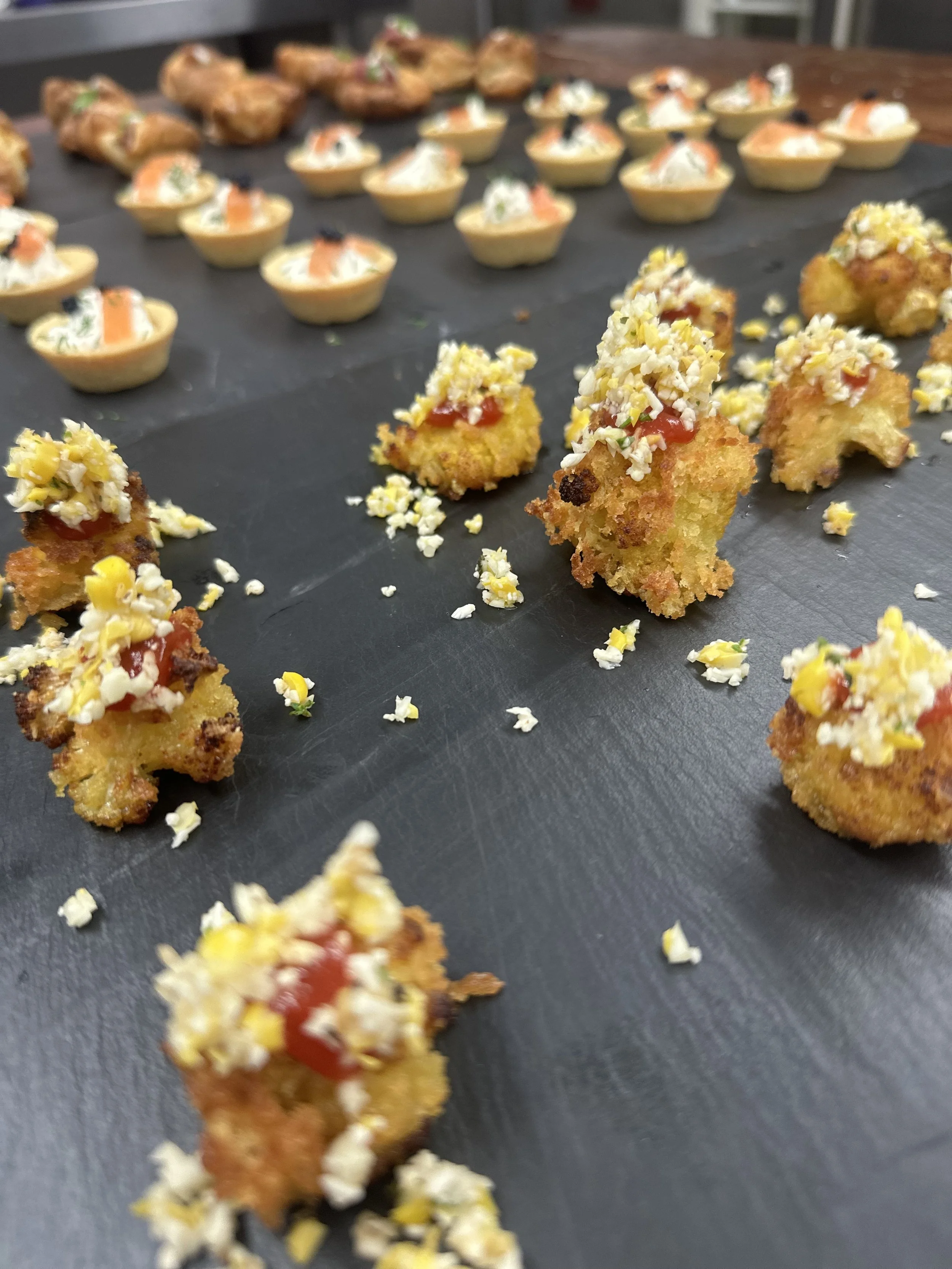 Close-up of bite-sized breaded appetizers topped with shredded cheese, chopped herbs, and a dollop of ketchup on a black serving board.