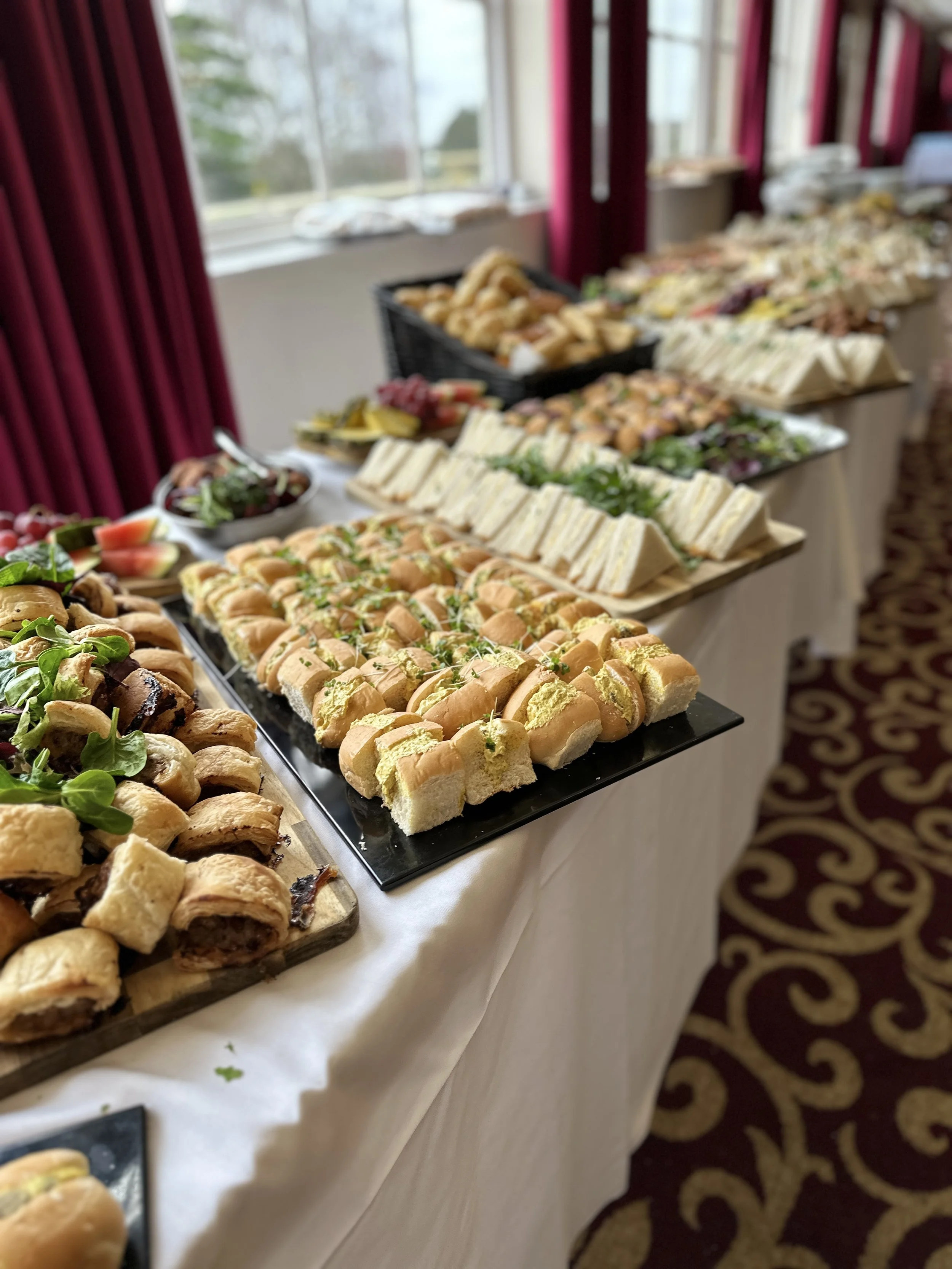 A buffet table with an assortment of sandwiches, mini hot dogs, pastries, and fresh fruit in a decorated room with burgundy curtains and patterned carpet.