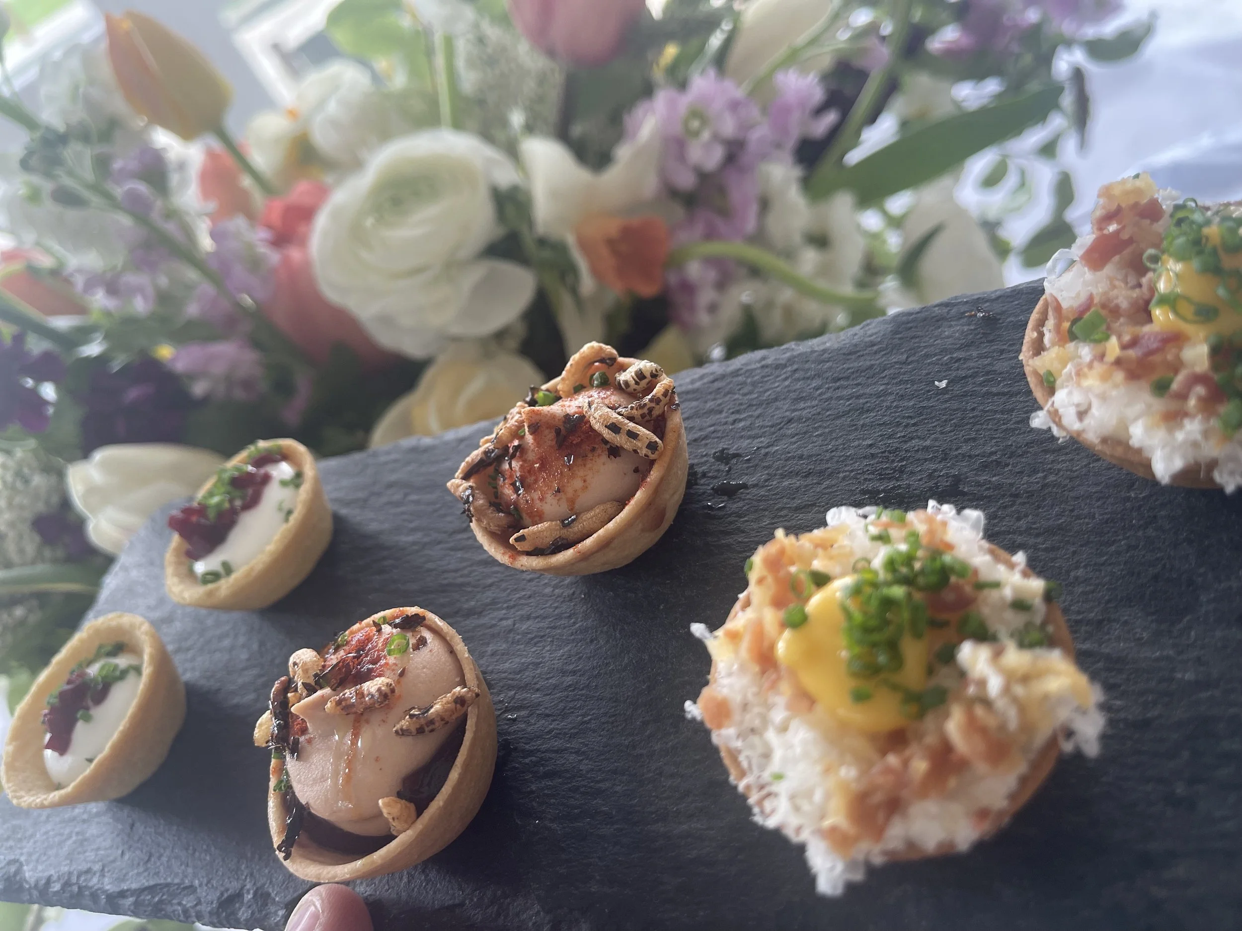 Assorted mini appetizers on a black slate serving platter with a floral bouquet in the background.