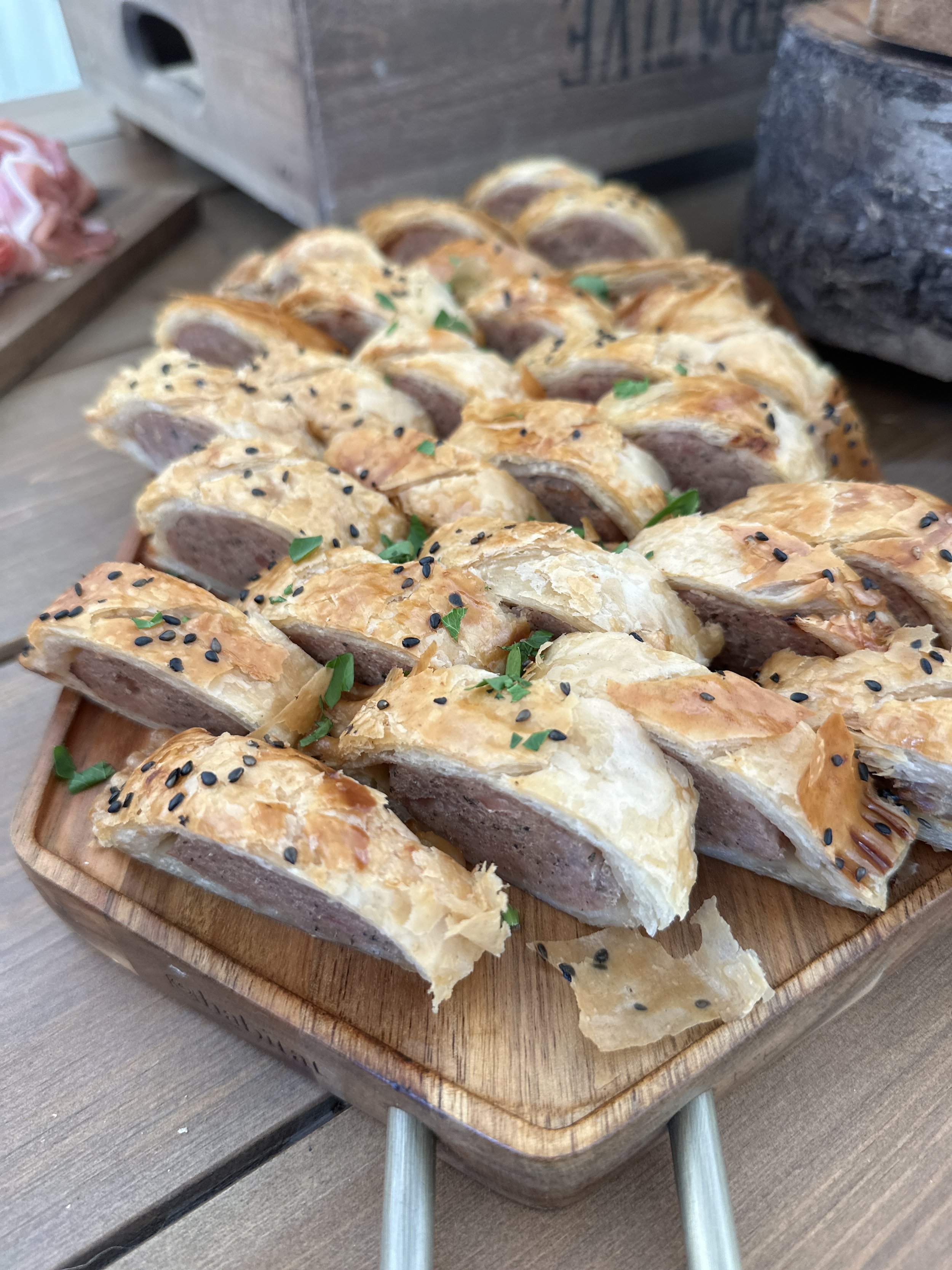 Slices of sausage in flaky pastry on a wooden serving tray, garnished with green herbs and black sesame seeds.