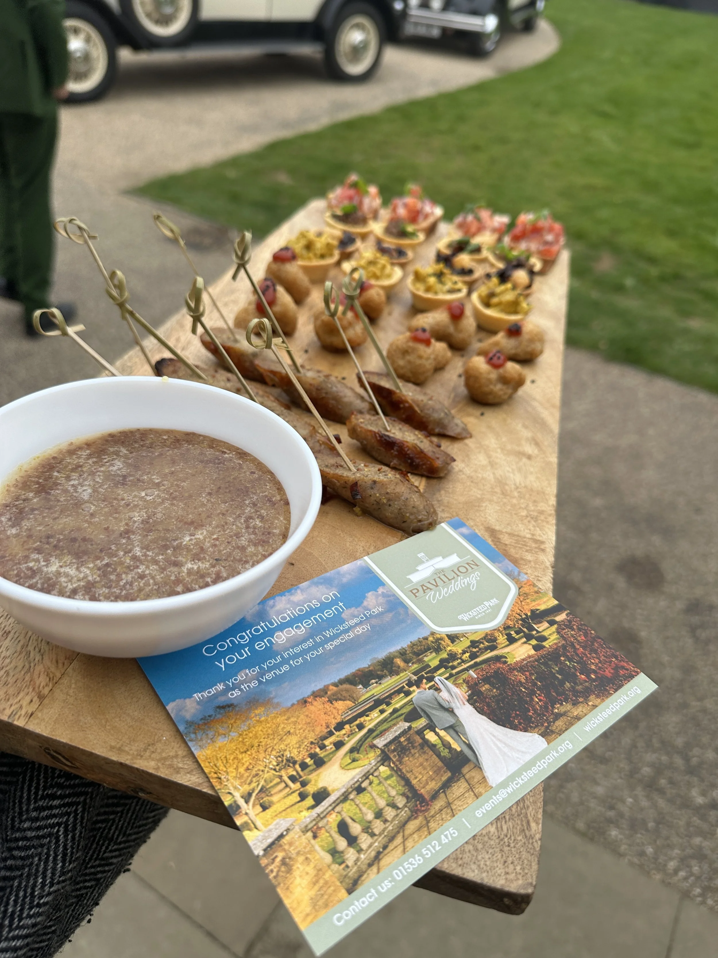 A wooden tray with small appetizers including meat skewers, mini tacos, and bite-sized snacks, a bowl of soup or sauce, and a wedding event flyer on a table outdoors.