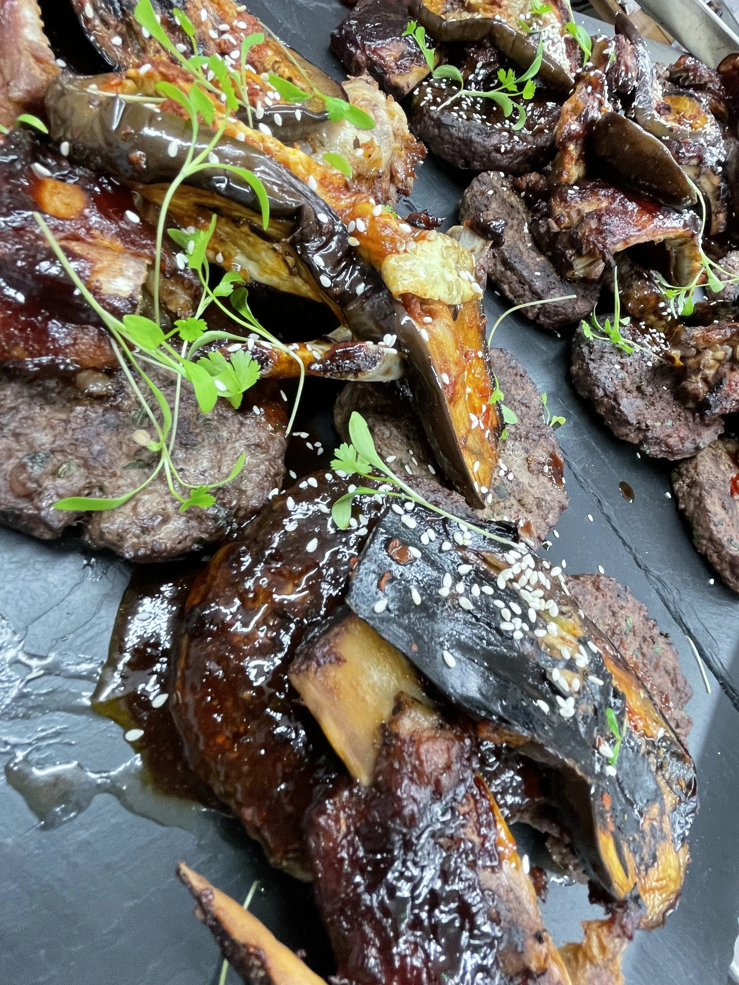 Grilled eggplant and beef patties garnished with sesame seeds and microgreens on a black serving tray.