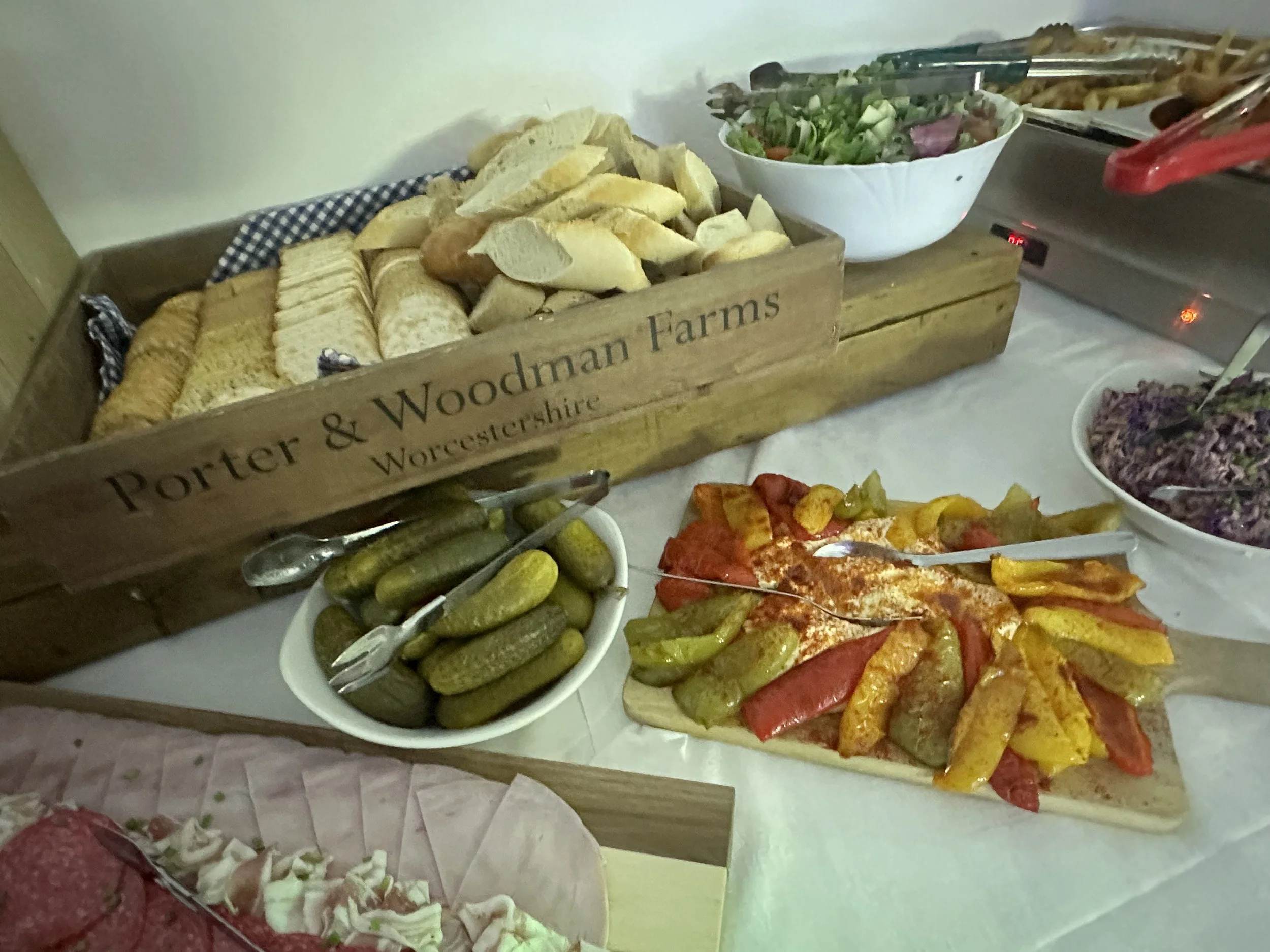 Variety of foods on a table, including a bowl of pickles, a pizza with roasted peppers, a salad, and a bread basket labeled 'Porter & Woodman Farms Worcestershire'.