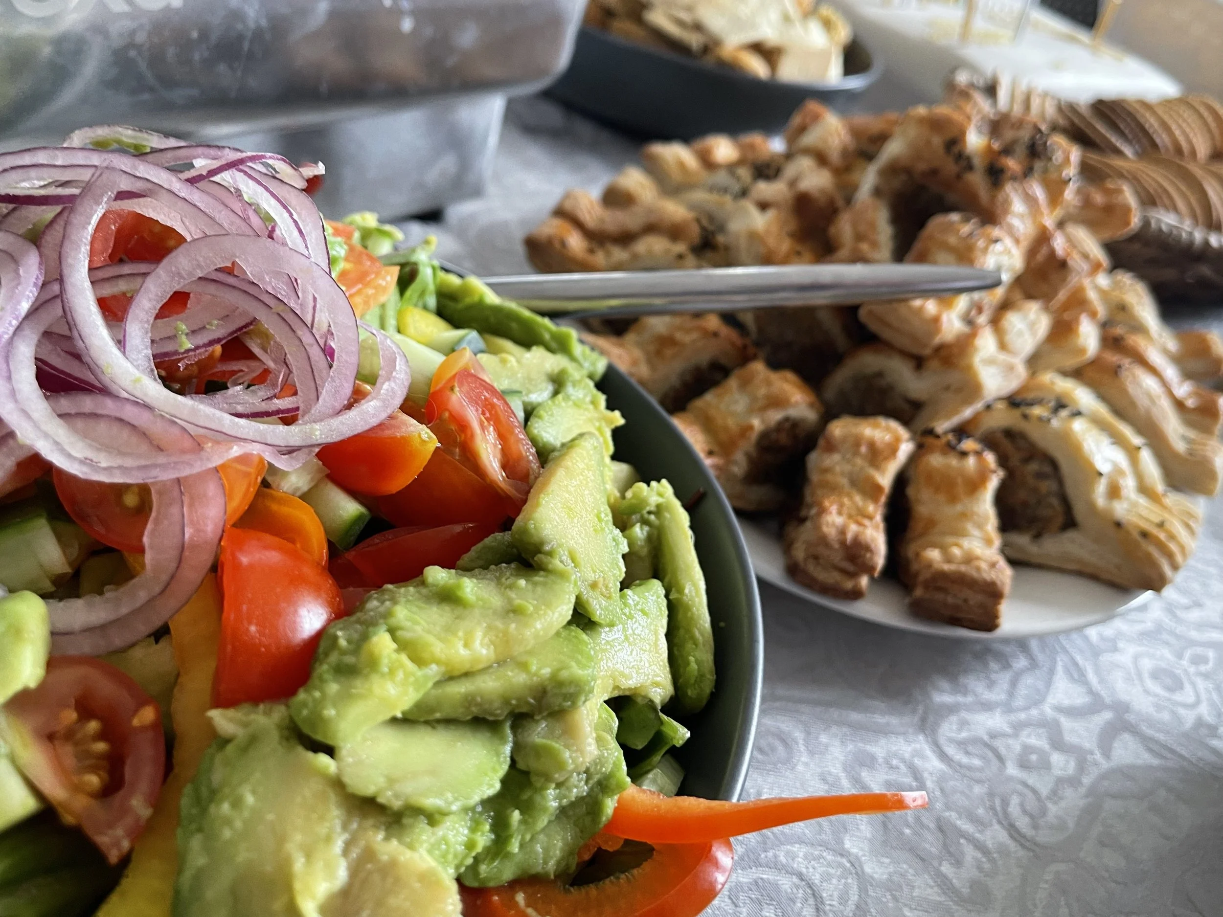 Close-up of a bowl of fresh vegetable salad with sliced onions, cherry tomatoes, avocado, and mixed greens, with a plate of assorted chicken and beef empanadas and other pastries in the background on a table.