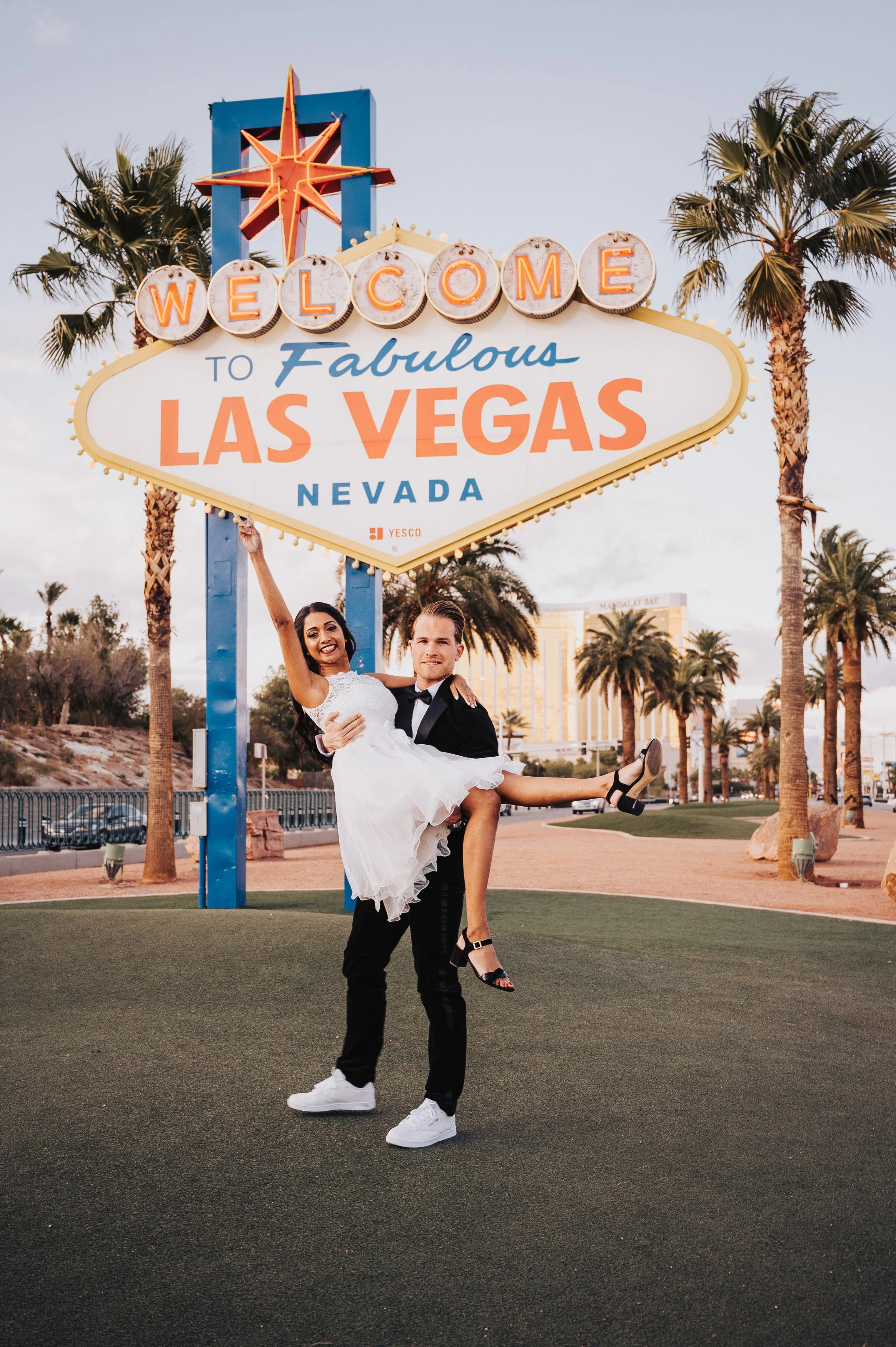 Ein Paar posiert glücklich vor dem „Welcome to Fabulous Las Vegas“ Schild. Der Mann hebt die Frau, die ein weißes Kleid trägt, hoch. Beide lächeln. Im Hintergrund sind Palmen und ein sonniger Himmel zu sehen.