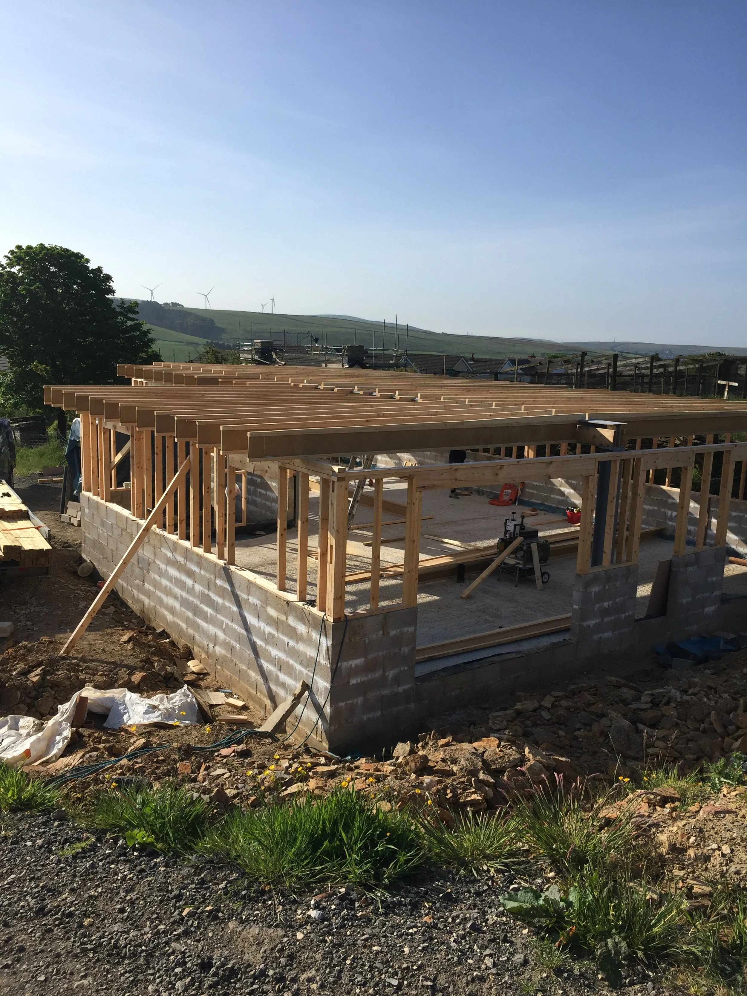 Construction site of a building with wooden framework and concrete block foundation, with a scenic landscape and wind turbines in the background.