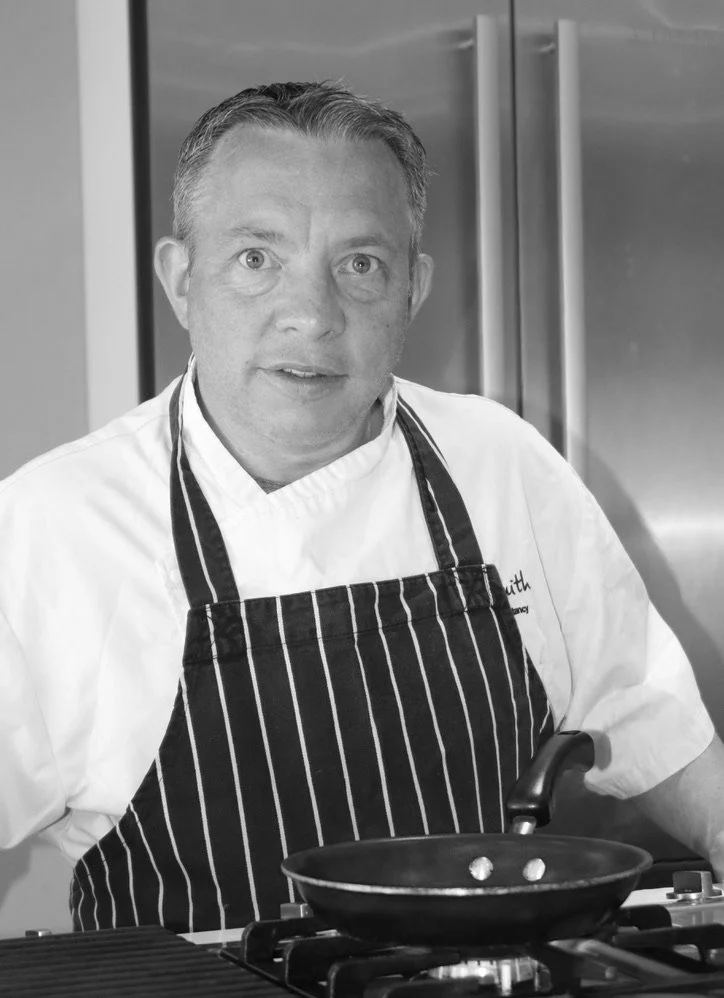 Chef wearing striped apron and white chef's coat standing by a stove with a frying pan.