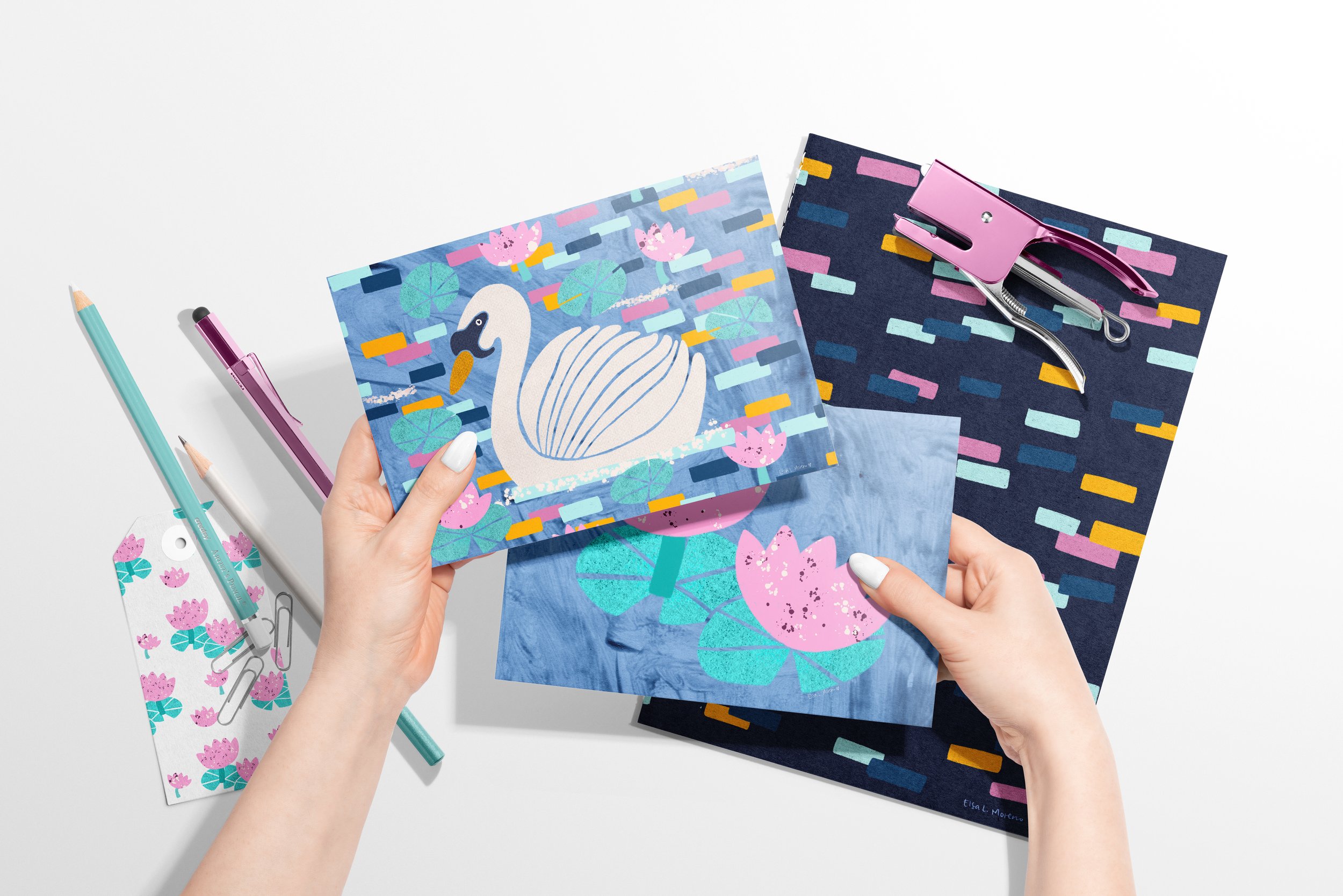 Two hands holding a card set of a swan and lily pads in pastel colors. The background shows several stationery articles, such as gift tags and paper board.