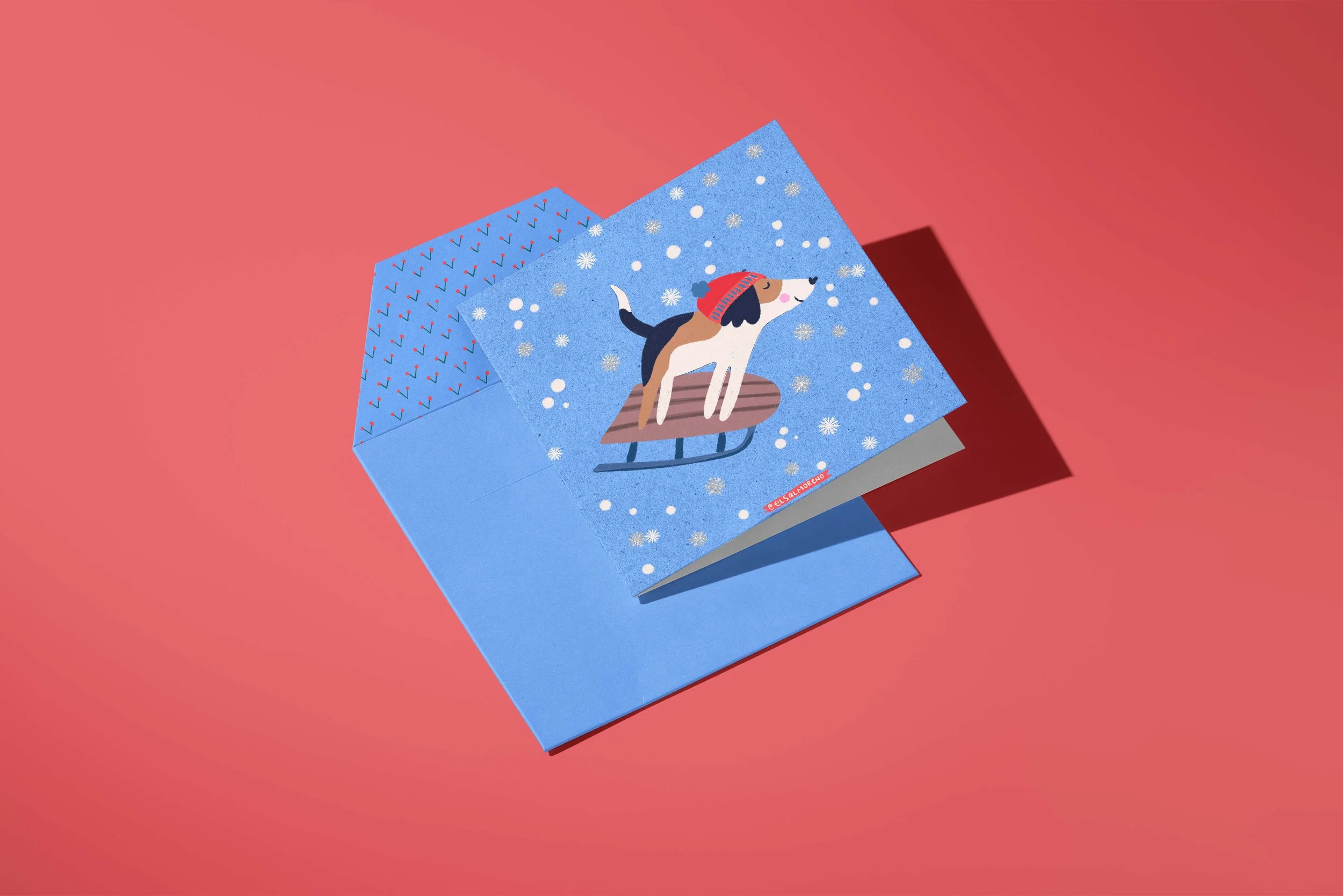 Square greeting card featuring a stylized dog sledding down with a smile on its face, surrounded by snow and snowflakes over a blue wintery sky.