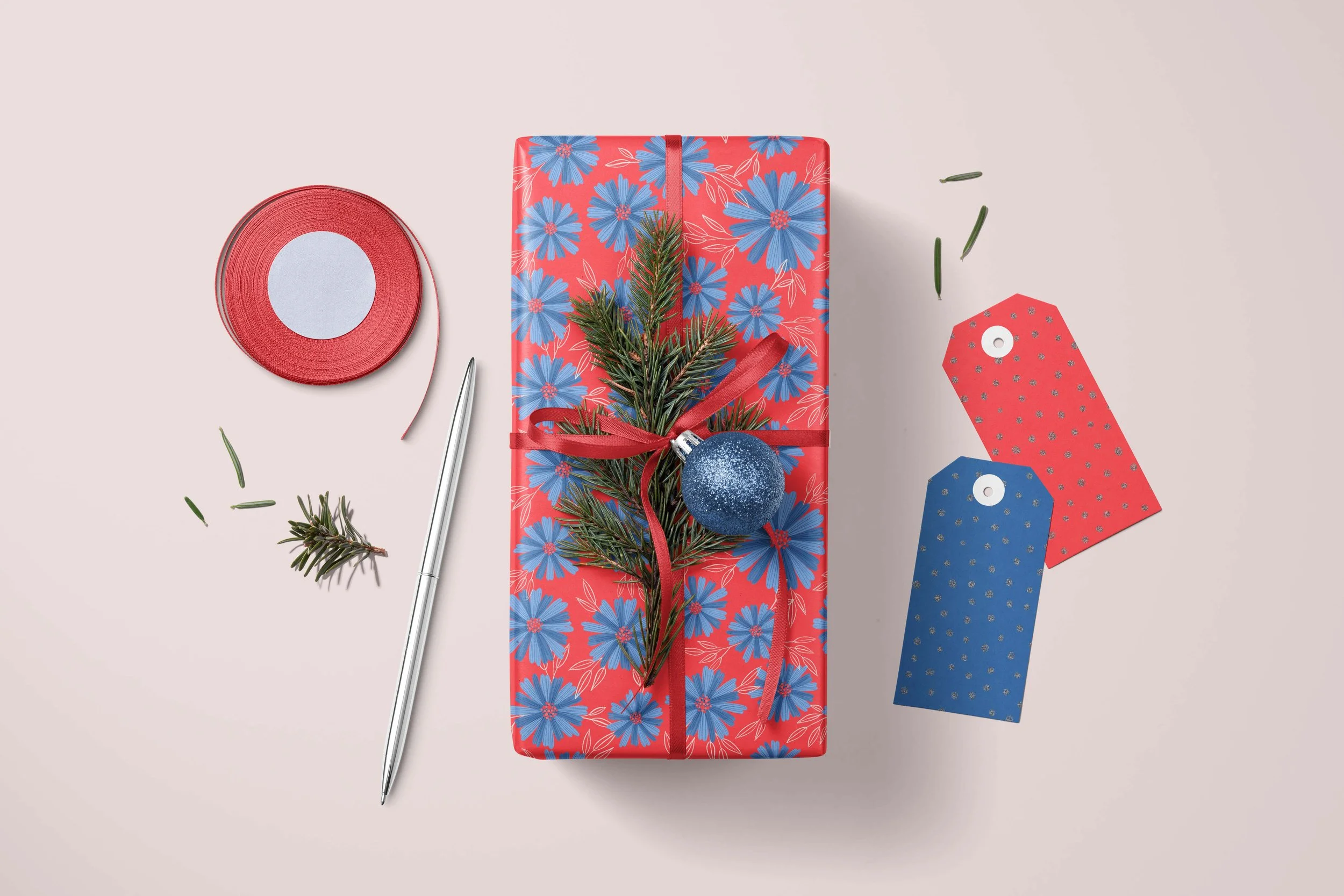 Picture of a Christmas gift wrapped in a cornflower and red floral motif, with a glittery blue tree ornament, red ribbon and pine branch. Around the gift there is some more red ribbon, a pen and two gift tags, one red, one blue.