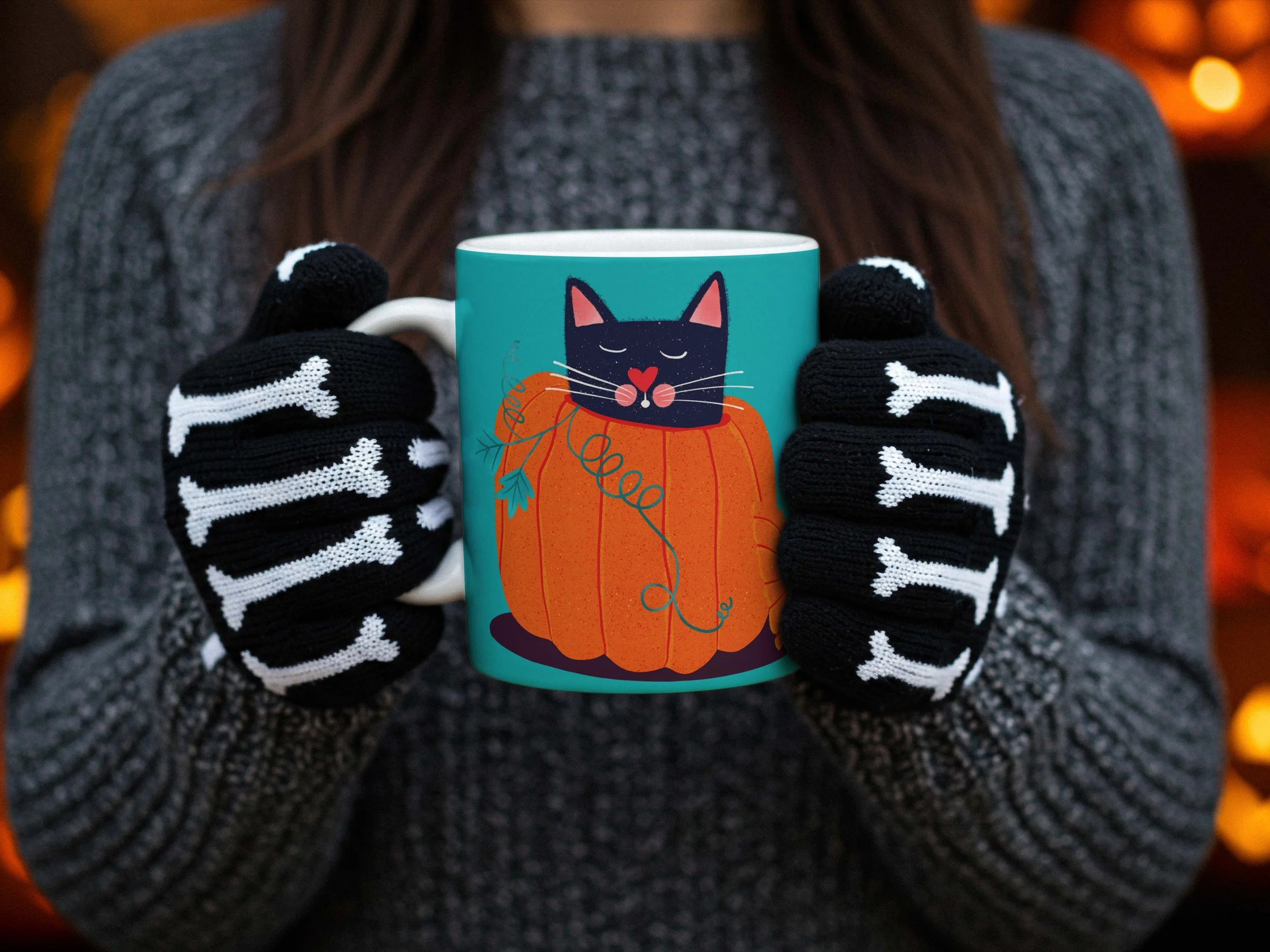 A woman wearing a grey knit sweater and black and white skeleton gloves holding a green Halloween mug depicting a black cat inside a pumpkin