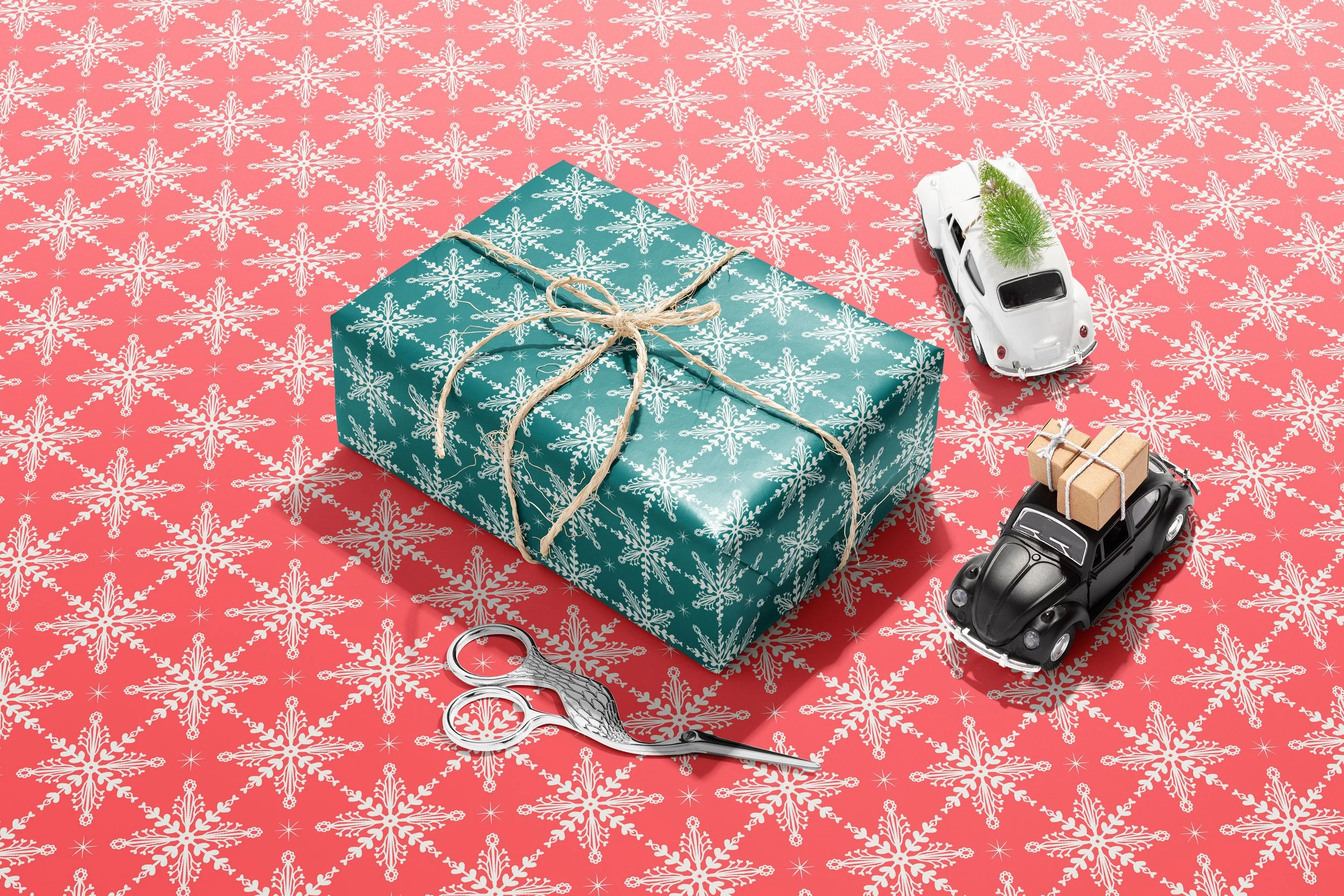 A present wrapped with white snowflake-motif over a green background wrapping paper on top of the same wrapping paper but with a red background. Two toy cars are beside the present, one white with a Christmas tree attached on top, one black with a co