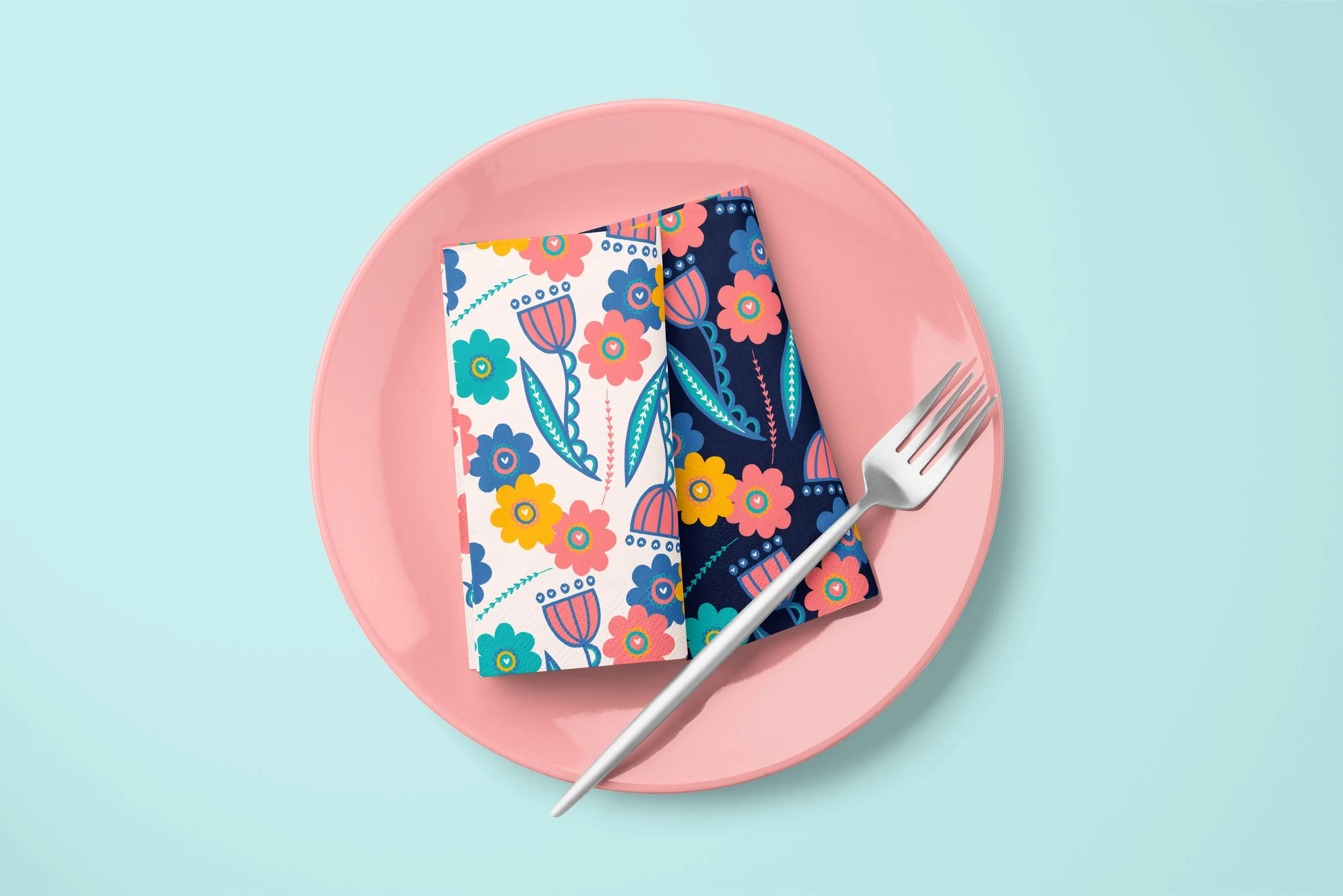 A coral-colored plate with two folded napkins depicting a floral pattern, one with light background and one with dark background
