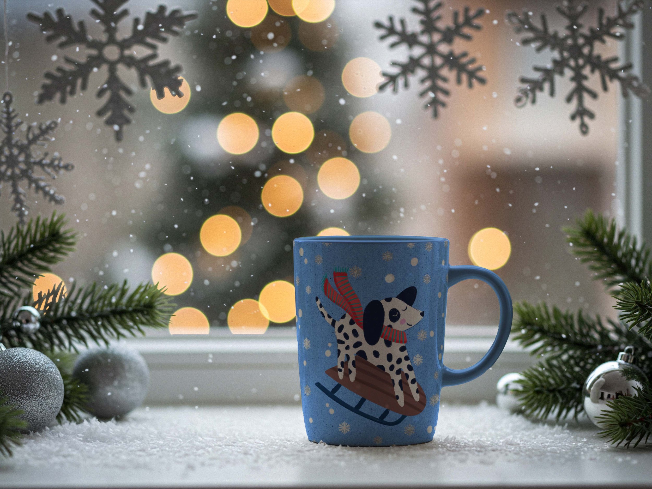 A cozy winter scene with a blue mug depicting a cute dalmatian in a red scarf sledding and surrounded by white snow and glittery silver snowflakes. The mug is on a white windowsill surrounded by hanging snowflakes, fir branches and silver Christmas o