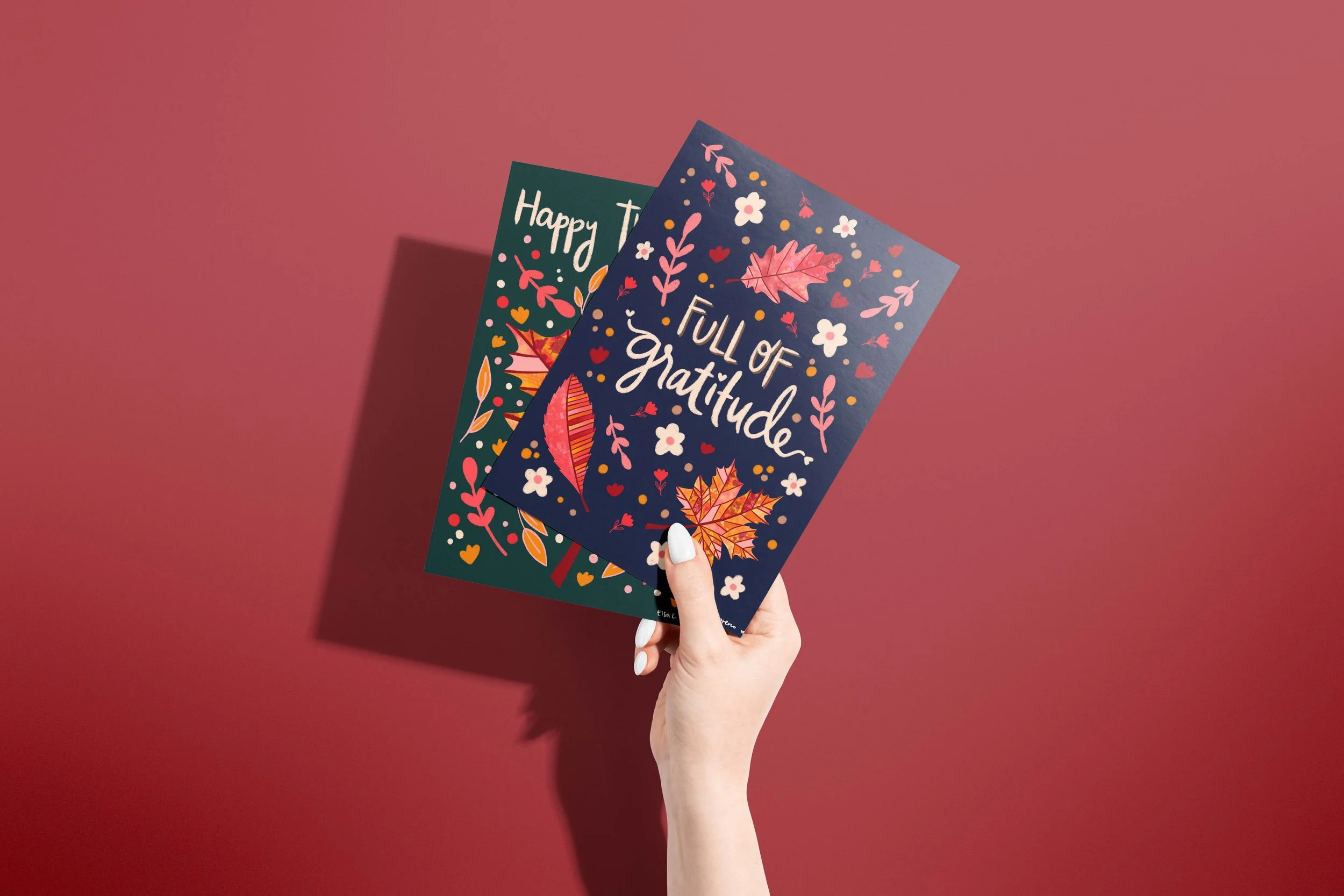 Two Thanksgiving cards featuring hand lettering and autumnal motifs, held by a woman's hand, over a burgundy background.