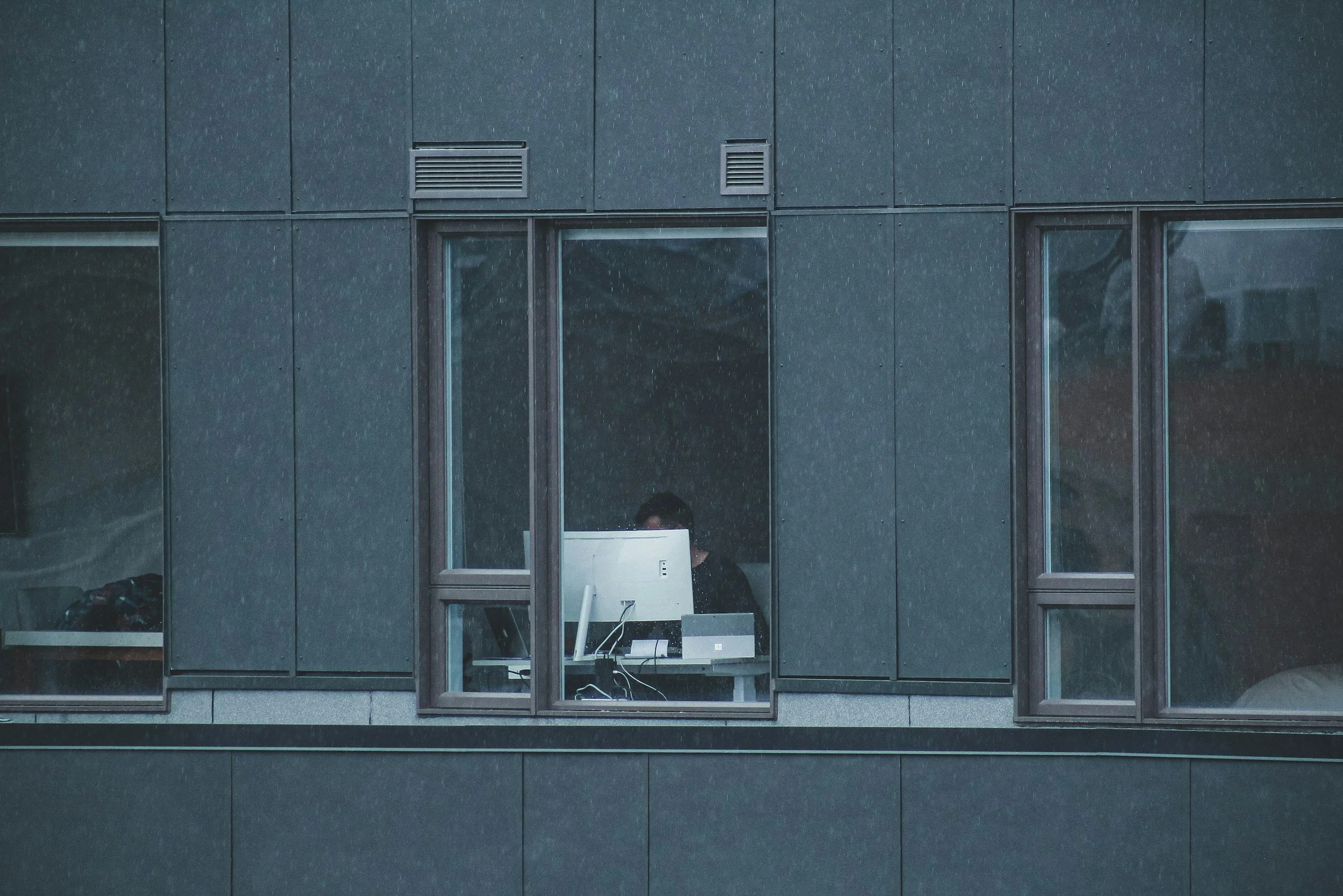 Person working at a desk with a computer in a modern building during rainy weather, visible through a window.