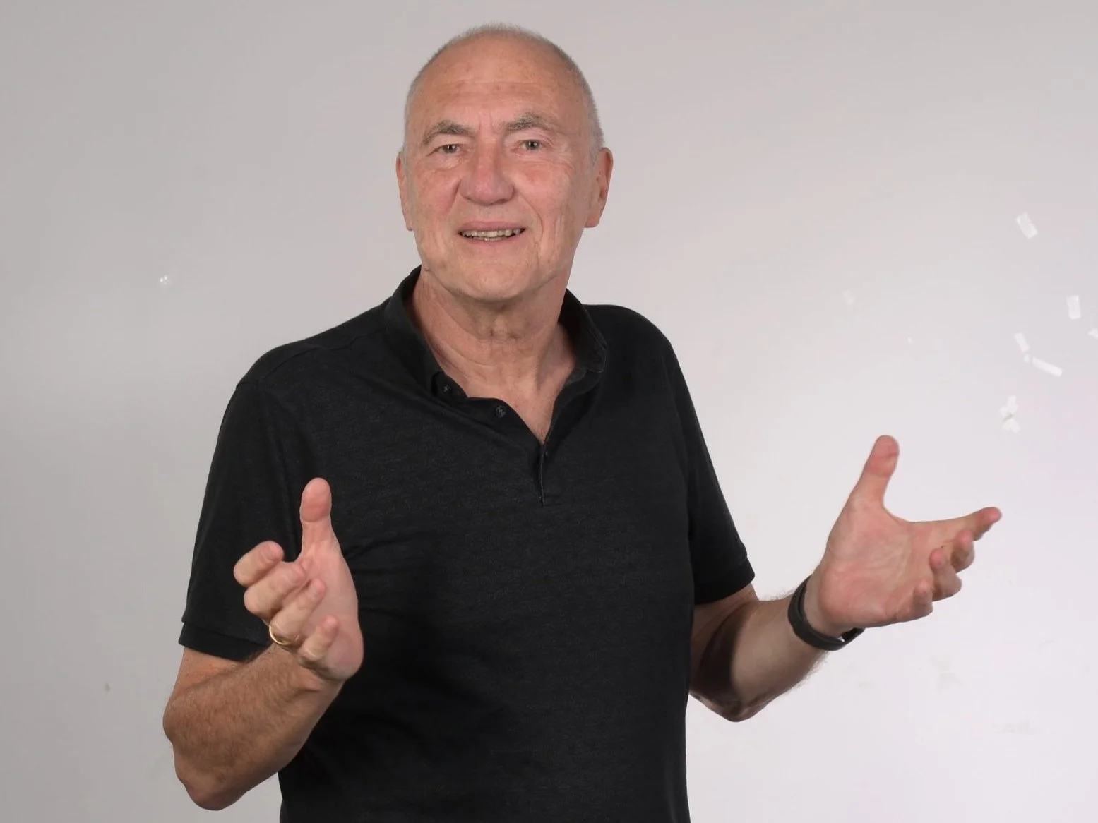 Older man in black shirt with expressive hands against a light background