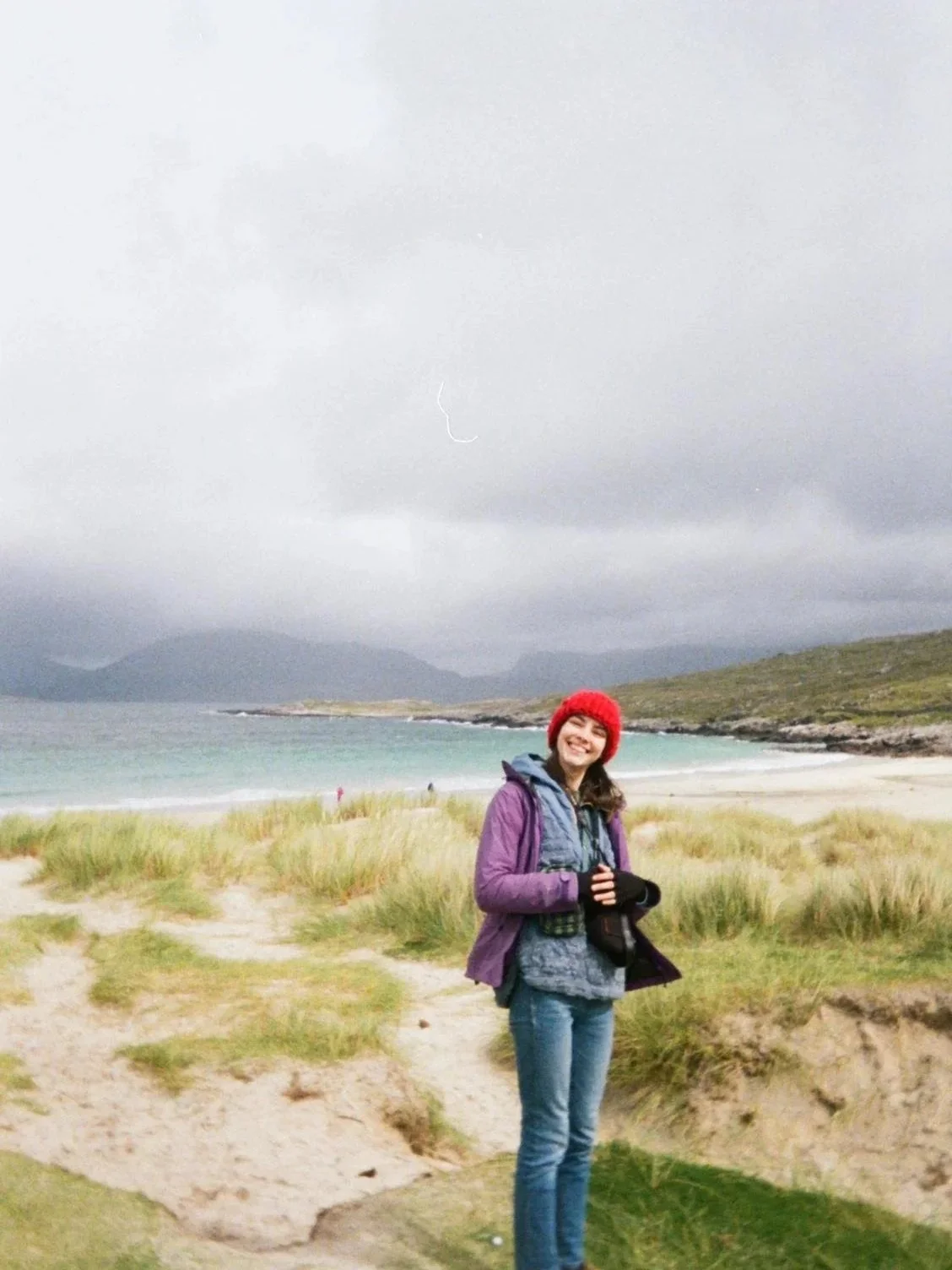A woman smiling at a beach with sand, grassy dunes, water, mountains, and overcast sky, dressed in a purple jacket, gray hoodie, red beanie, and gloves.