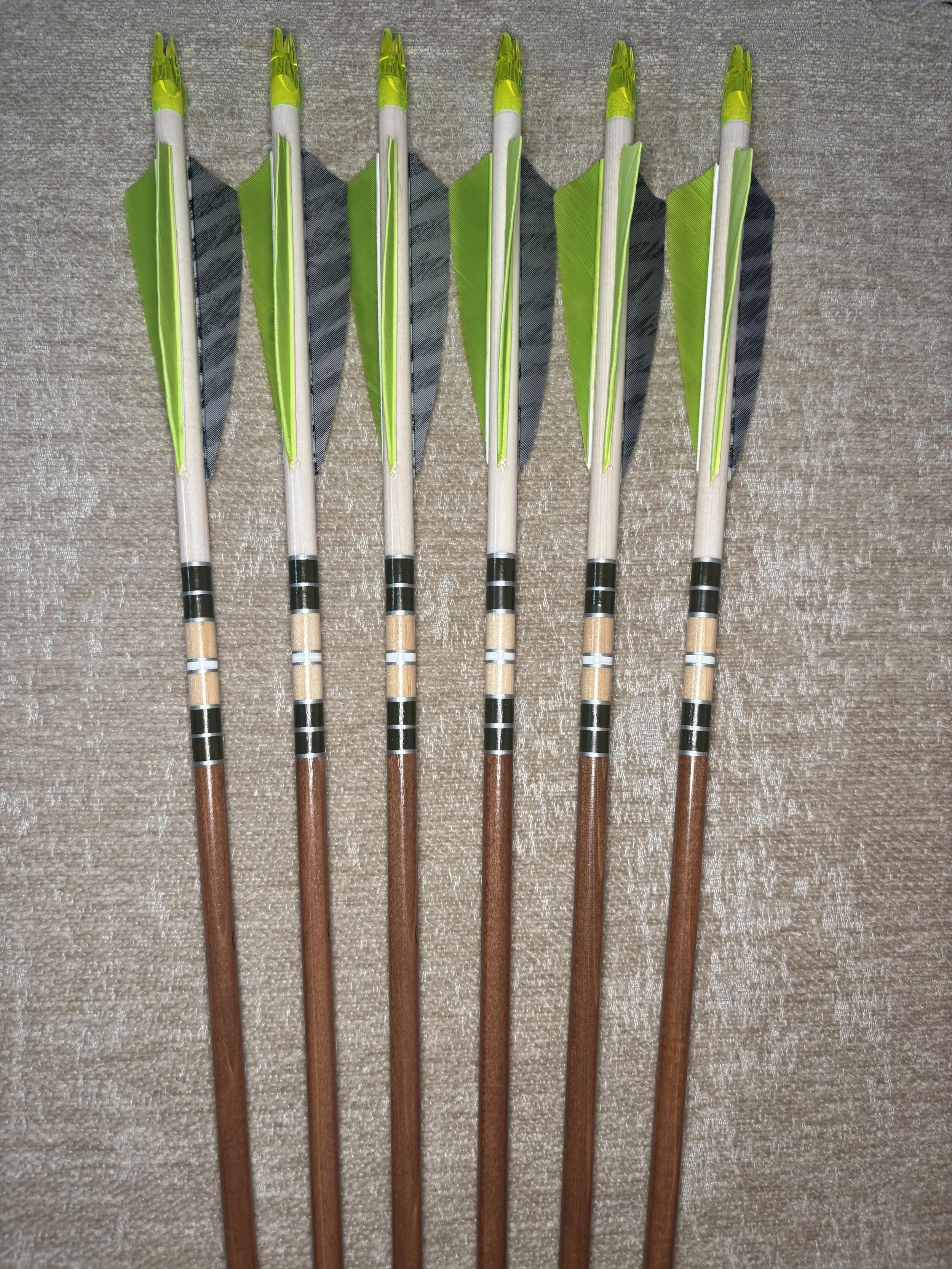 Six archery arrows with green fletching and brown shafts, arranged parallel on a beige carpet.