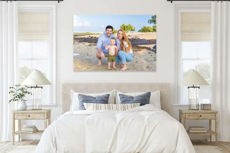 A large framed family photo of a man, woman, and young boy on the beach hangs above a white bed in a bright bedroom with white walls, two windows, and matching nightstands with lamps.