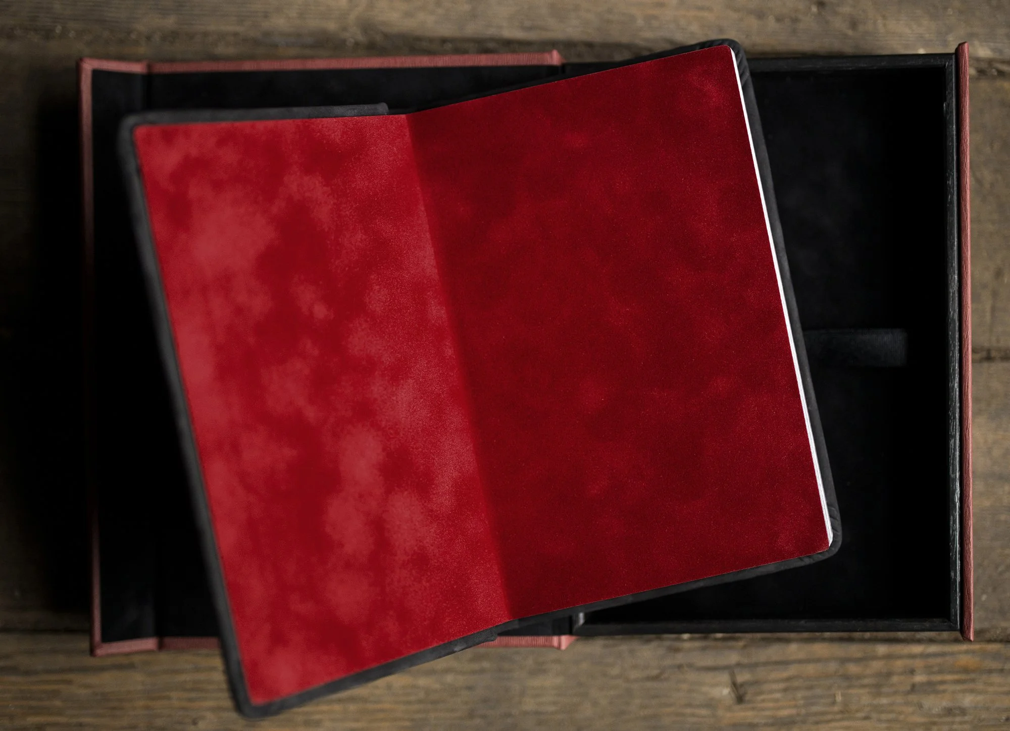 Open red leather-bound book or album with black edges, placed inside a black box, on a wooden surface.