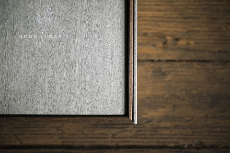 Close-up of a corner of a gray textured panel with a logo of a leaf and the words 'anna maria' on it, placed on a wooden surface.