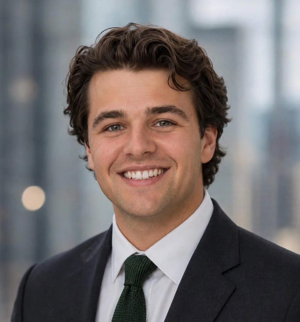 A light-skinned person smiling at the camera with short, dark, curly hair, wearing a dark suit.