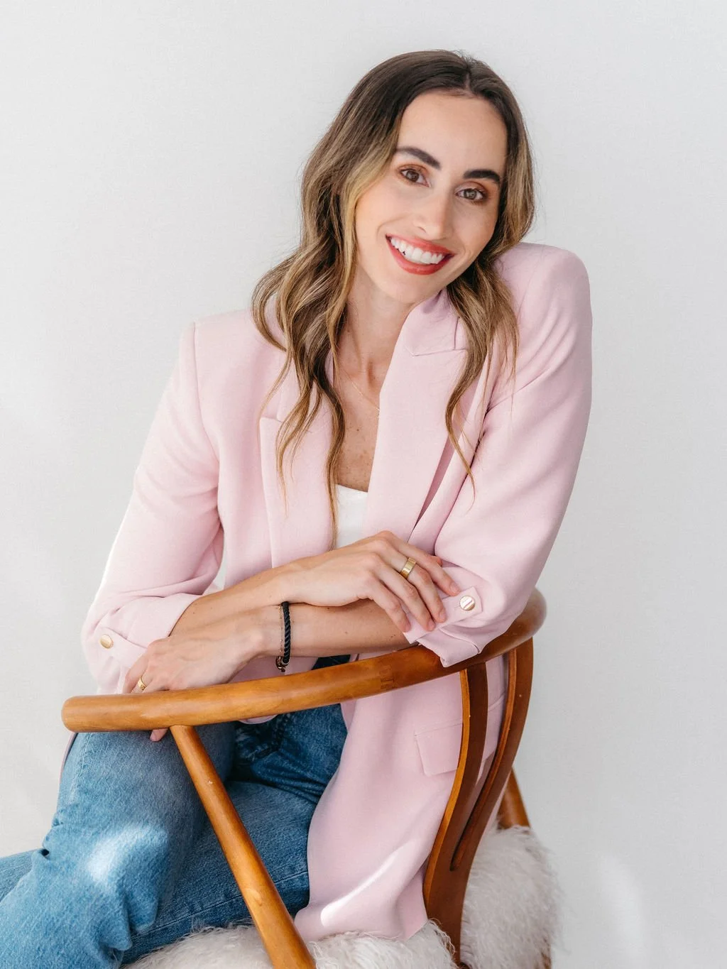 A woman with wavy brown hair, smiling, dressed in a light pink blazer and white top, sitting on a wooden chair with a fluffy white cushion, against a plain white background.