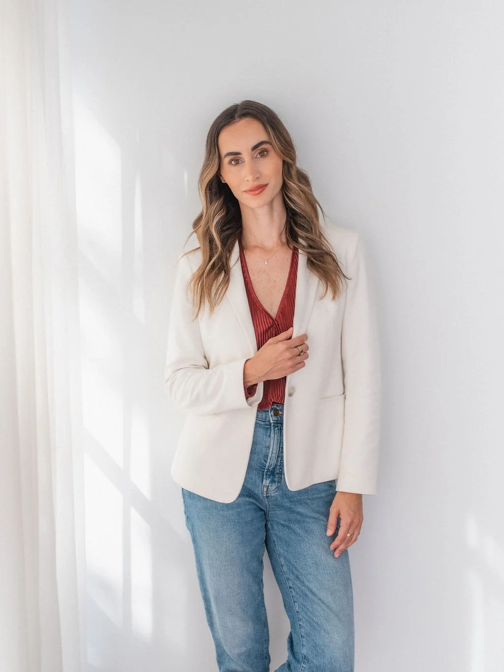 Woman with wavy brown hair wearing a white blazer, red blouse, and blue jeans standing against a white wall.