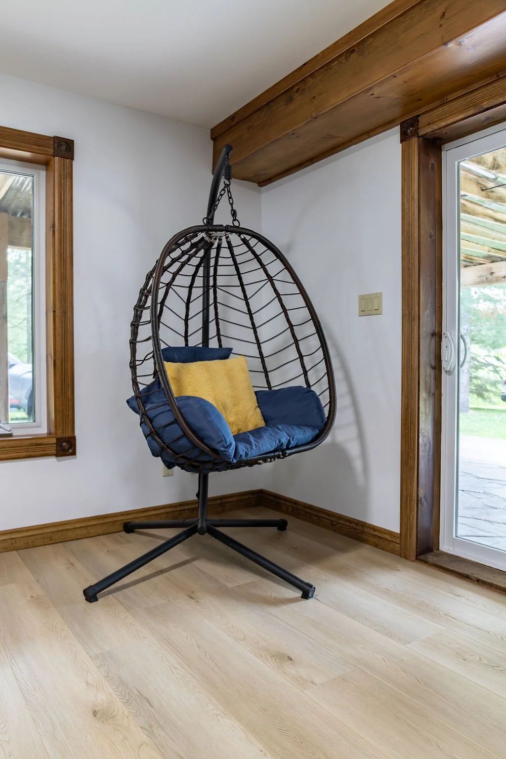 A hanging outdoor-style hanging egg chair with a black metal frame and brown rattan exterior, featuring blue cushions and a yellow throw pillow inside, situated in front of a sliding glass door and a window in a room with wooden trim and light wood f