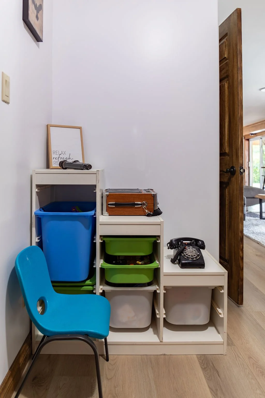 A white shelving unit with various storage bins, a vintage black rotary phone, a framed quote, and a blue child's chair in front of a white wall, next to a wooden door opening to a living room.