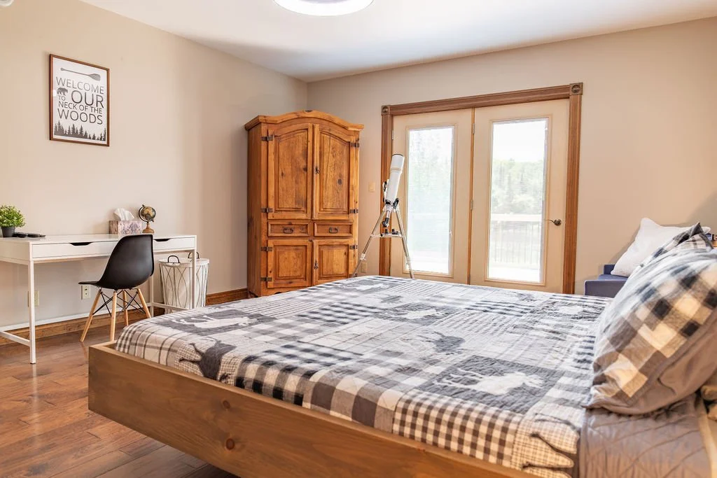 A bedroom with a wooden bed, checkered bedding, and pillows. There's a wooden armoire, telescope, sliding glass door opening to a balcony, desk with a chair, and wall art that reads "Welcome to our neck of the woods."