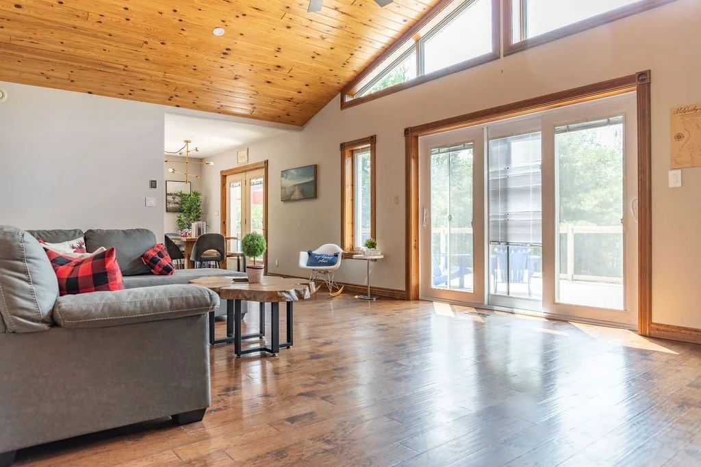 Living room with a gray sofa, plaid pillows, a wooden coffee table, a dining area, and large sliding glass doors leading to an outdoor balcony with trees visible outside.