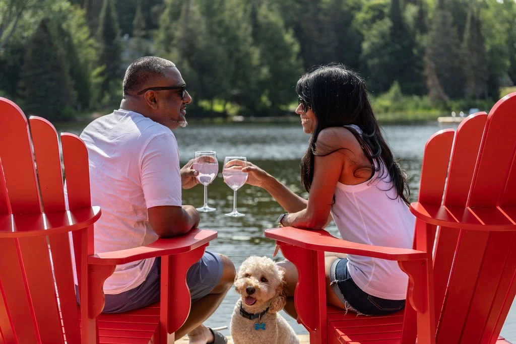 A couple sitting by a lake on red Adirondack chairs, toasting glasses of water, with a dog sitting between them.