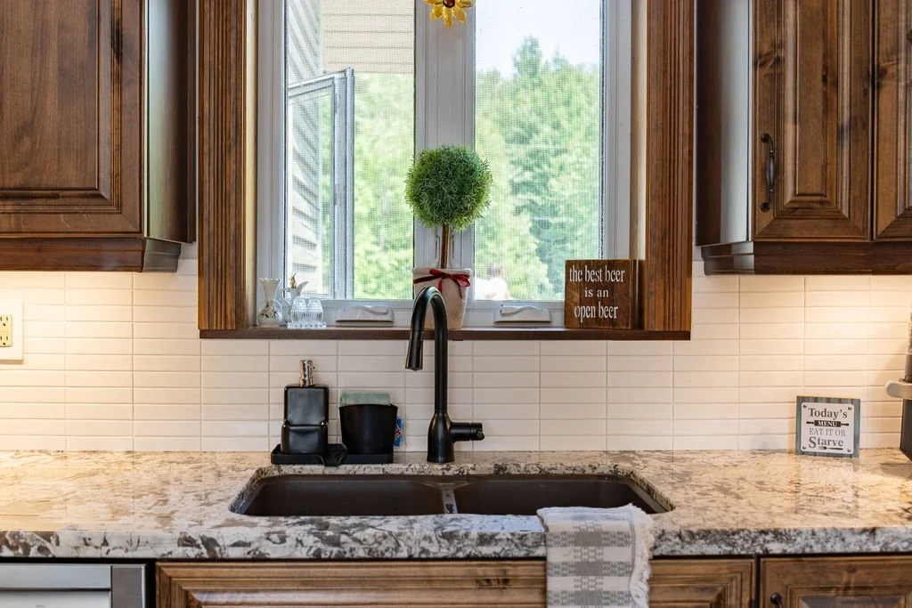 Kitchen sink with black faucet, soap dispenser, and sponge, set in a granite countertop below a window with a small potted plant and a decor sign saying 'the best beer is an open beer,' surrounded by wooden cabinets.