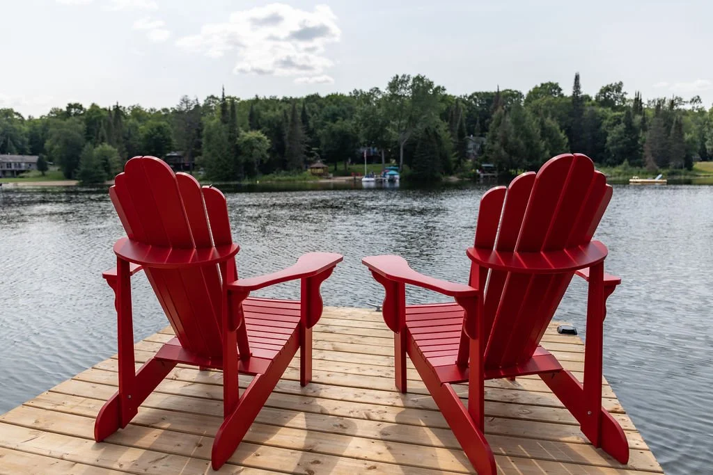 Two red Adirondack chairs on a wooden dock by a lake, with trees and houses in the background.