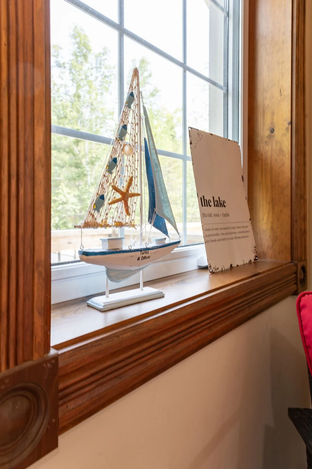 Decorative model sailboat on a wooden windowsill next to an informational sign titled 'the lake' in a room with wooden trim and a screen window.