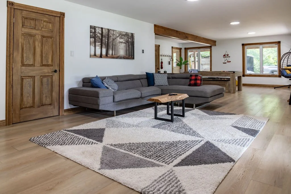 Spacious living room with a gray sofa, decorative pillows, a geometric patterned rug, a wooden coffee table, large windows, and wooden accents.