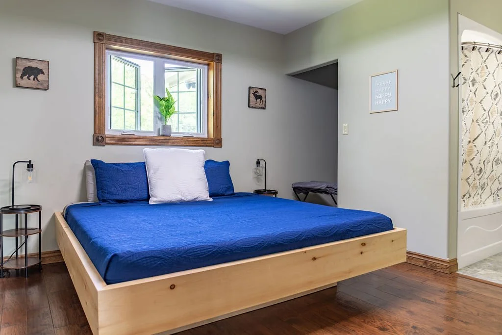 Bedroom with a wooden bed frame, blue bedding, and a white pillow, next to two small black nightstands with lamps, a window with a potted plant, and wall art that reads 'Happy'.