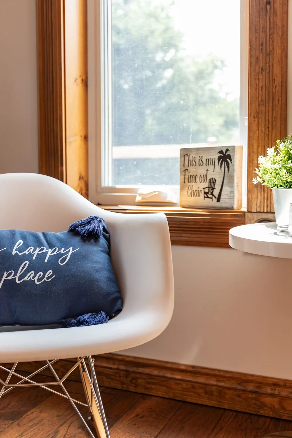 A cozy corner with a white modern chair featuring a blue cushion that reads 'happy place,' positioned near a window with wooden trim. On the window sill, there is a sign that says 'This is my time out chair' with a palm tree and chair illustration, a